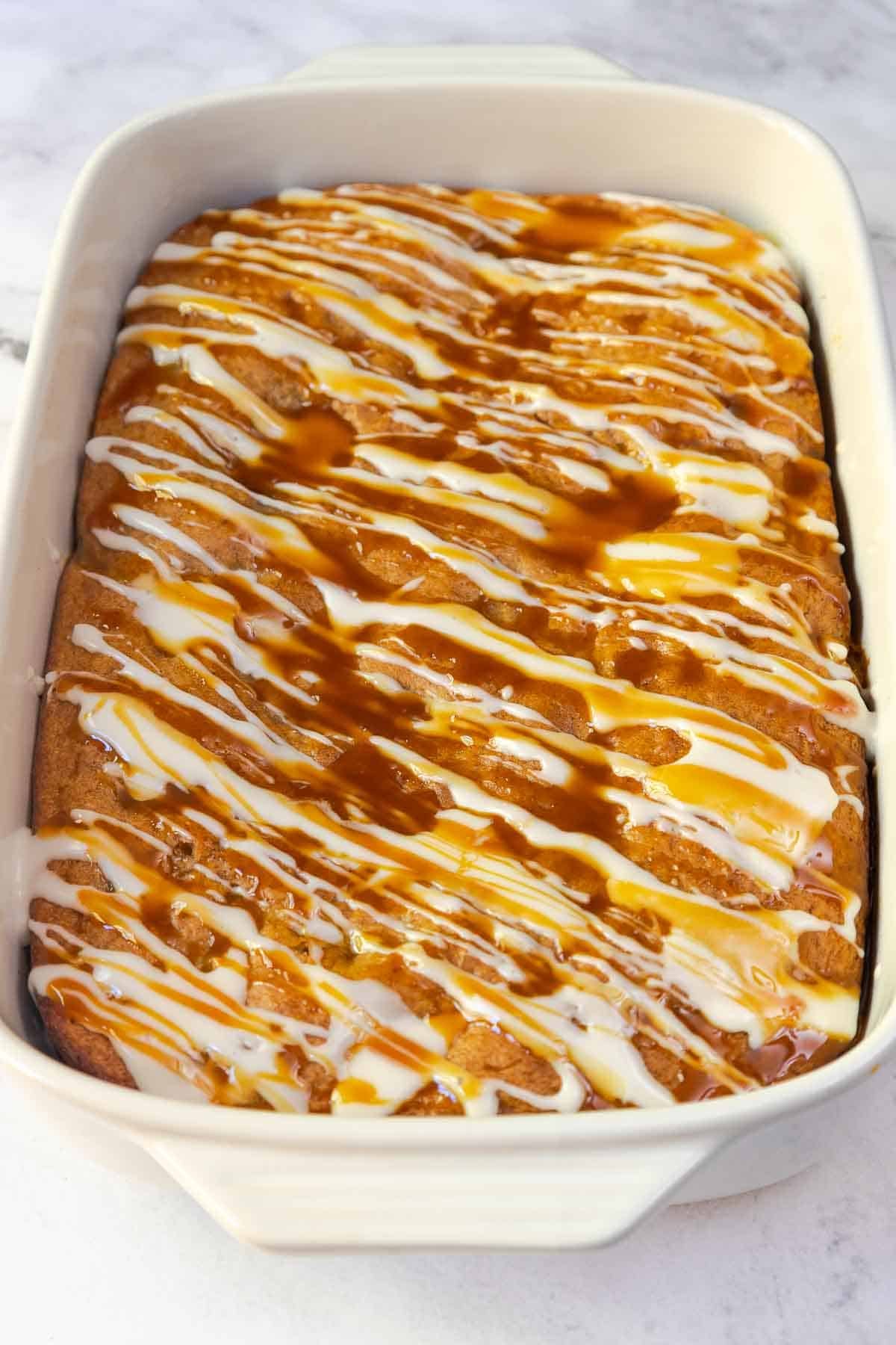 The finished apple cake in a 9 by 13 inch baking dish, topped with a generous drizzle of both white glaze and caramel sauce.