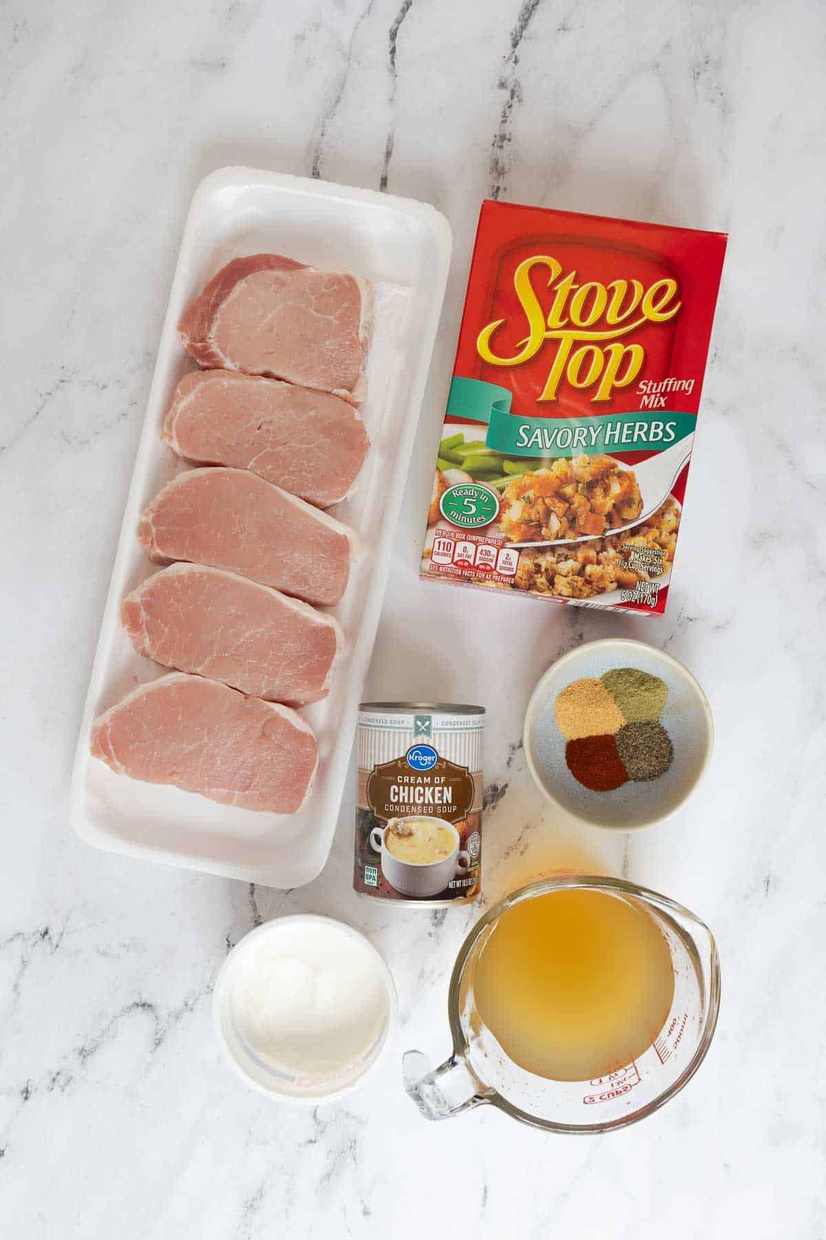 Ingredients for baked pork chops with stuffing including raw pork chops, Stove Top stuffing mix, cream of chicken soup, sour cream, chicken broth, and a bowl of mixed spices.