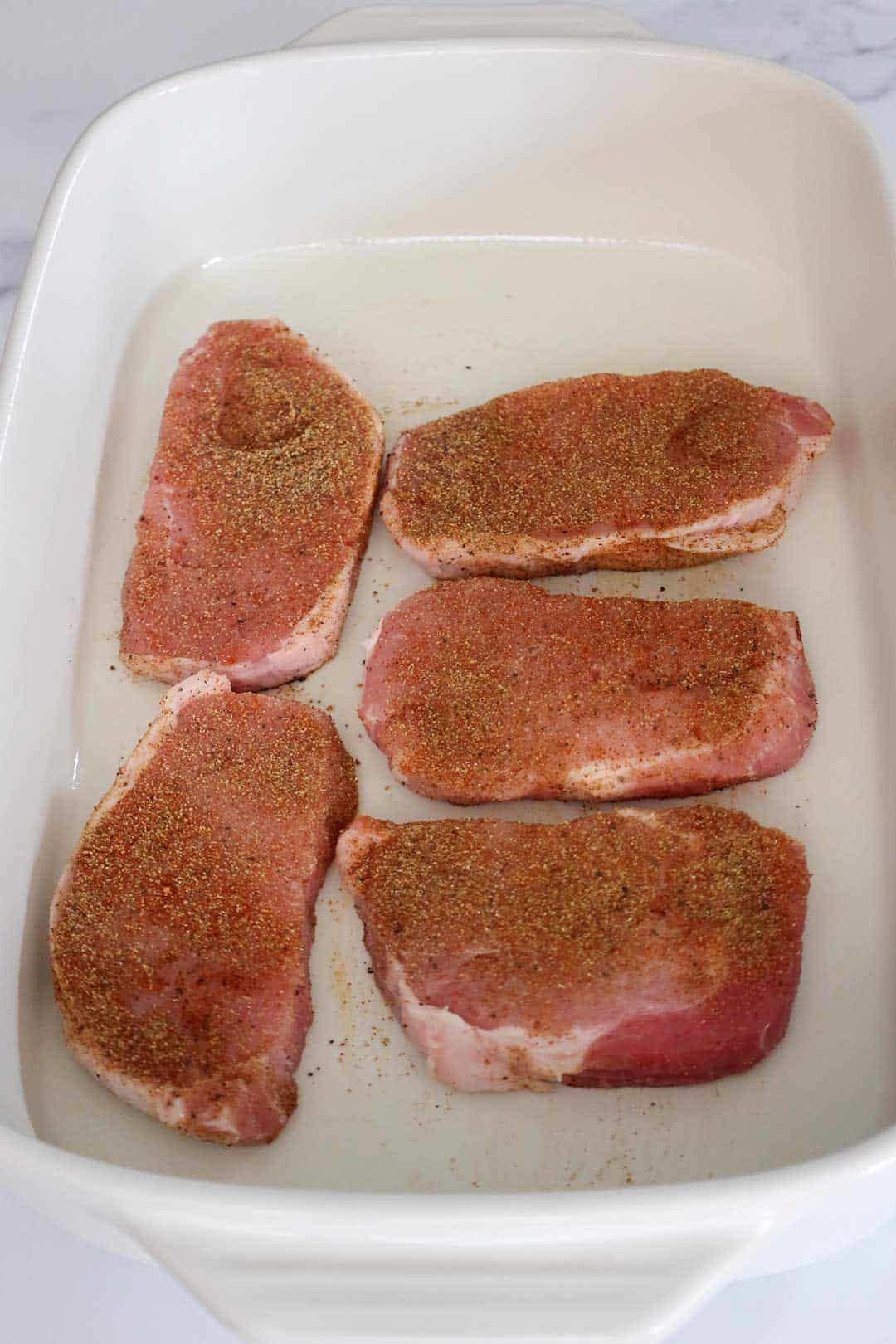 Five seasoned raw pork chops arranged in a white baking dish, ready for the next step in the recipe.