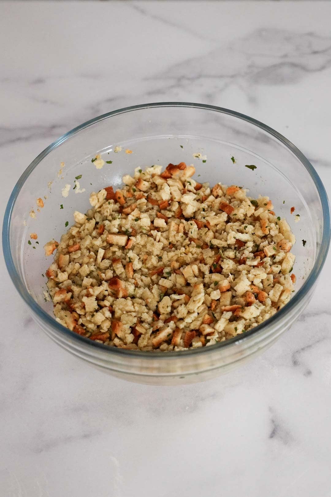 A glass bowl filled with prepared stuffing mix, showing cubed bread pieces and dried herbs.