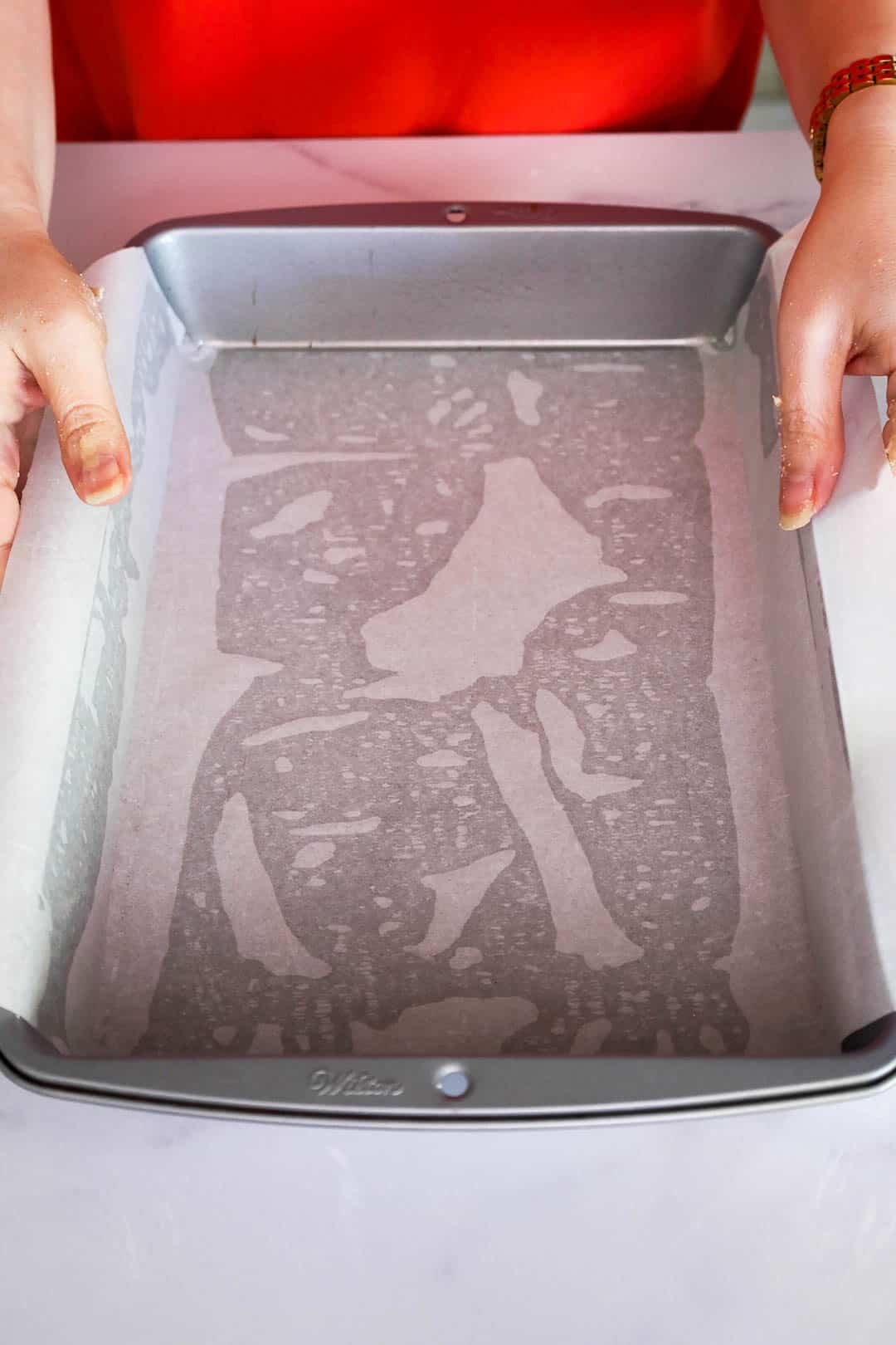A parchment-lined metal baking pan being prepared by hand, ready to be filled with cookie dough.