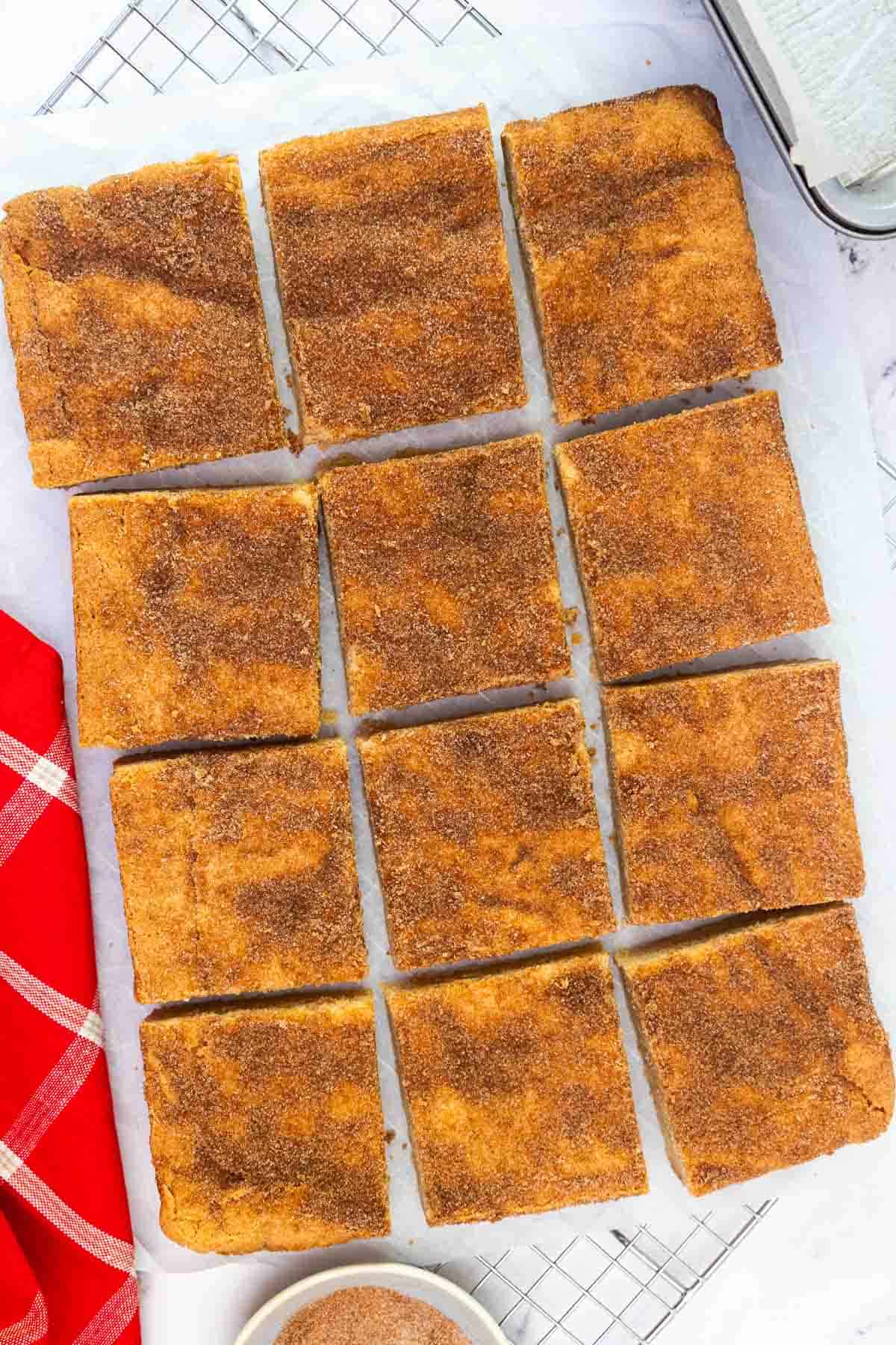 Twelve evenly cut snickerdoodle cookie bars arranged on parchment paper over a cooling rack, ready to serve.