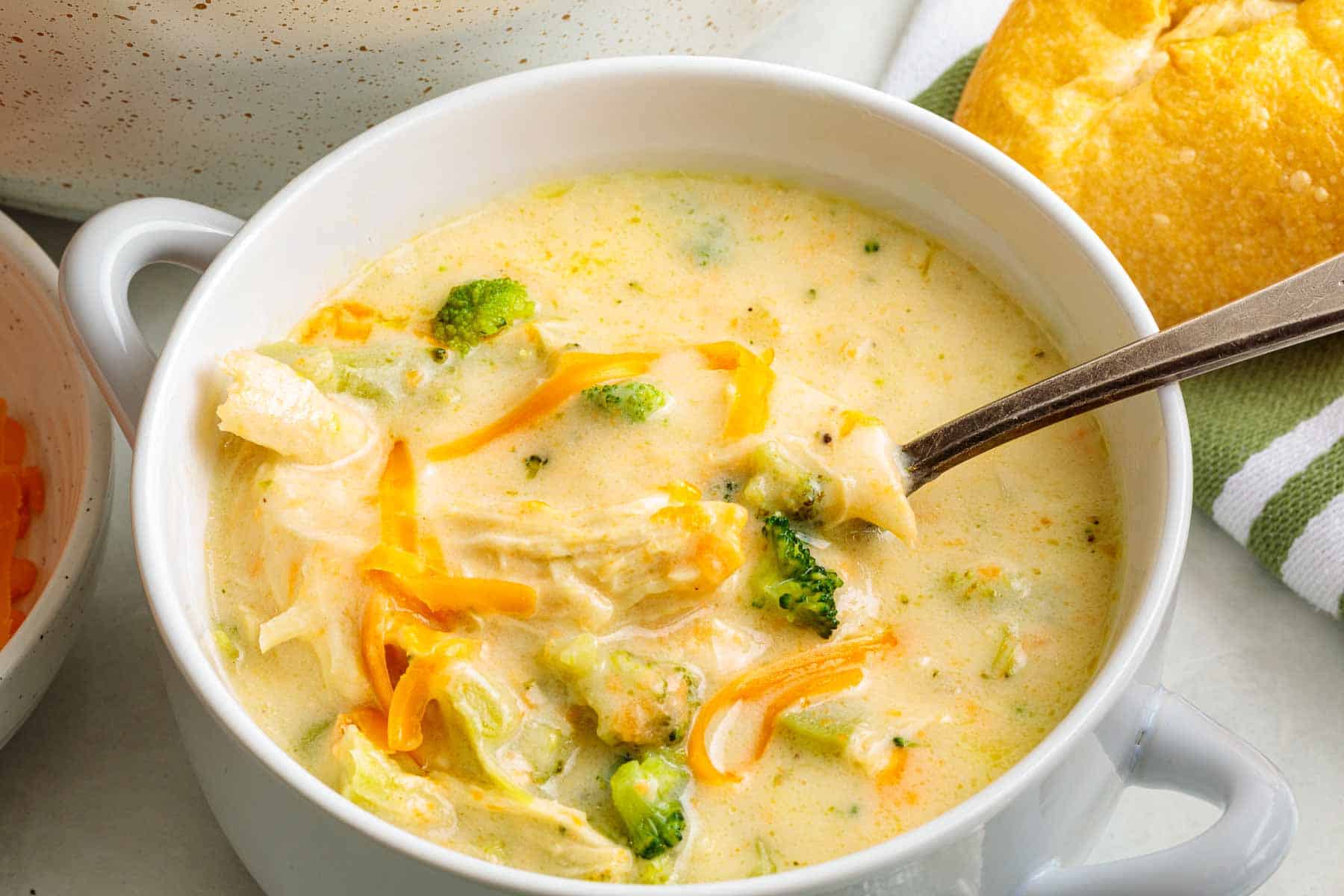A bowl of chicken broccoli cheddar soup with a spoon resting inside, revealing chunks of chicken, broccoli florets, and cheddar cheese in a creamy base.