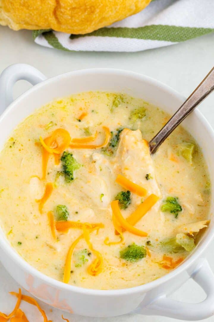 Overhead view of a bowl filled with chicken broccoli cheddar soup, garnished with shredded cheddar and surrounded by a crusty roll and napkin.