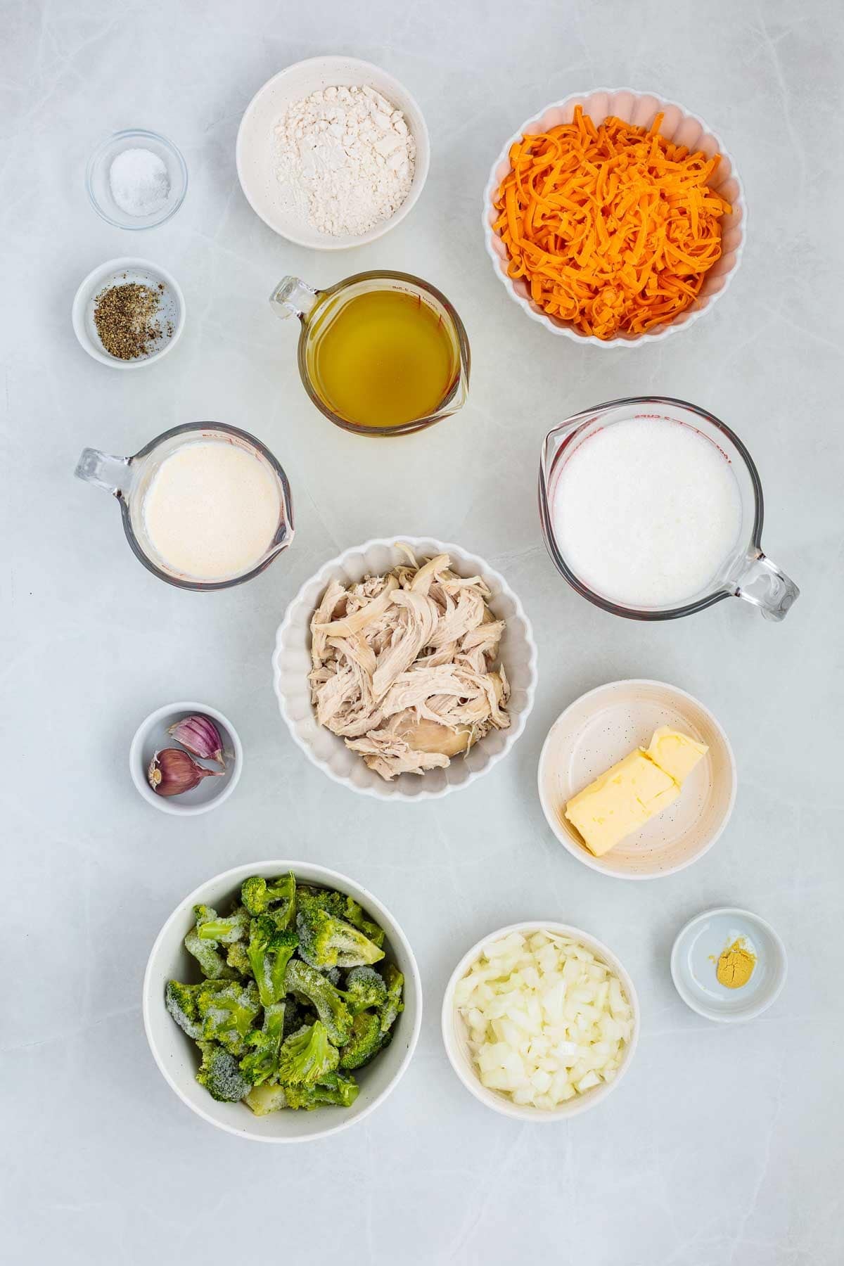 Flat lay of the soup ingredients including shredded cheddar, cooked chicken, frozen broccoli, chopped onions, garlic cloves, flour, butter, broth, cream, milk, mustard powder, salt, and pepper.