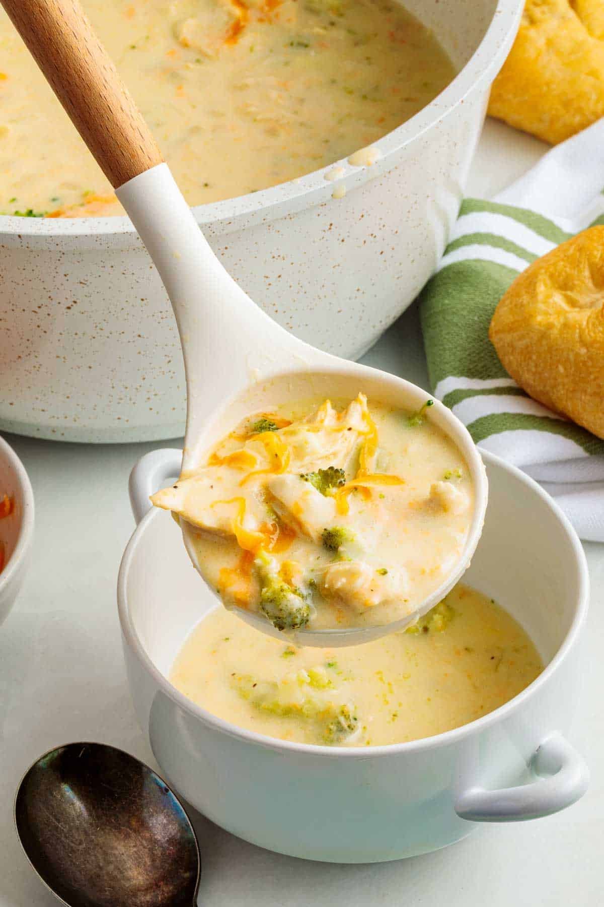 A ladle filled with steaming chicken broccoli cheddar soup being served into a white bowl, with visible chunks of chicken, broccoli, and melted cheddar.