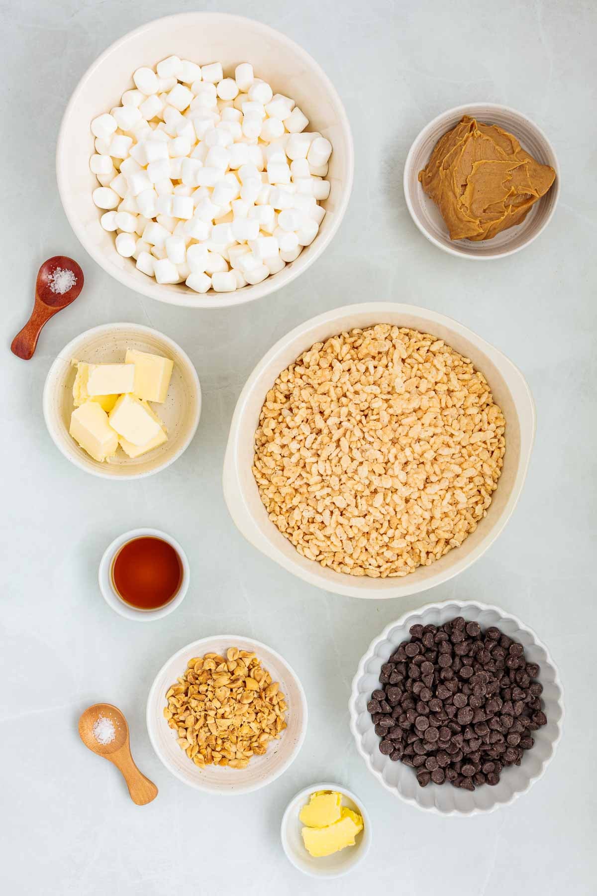 Flat lay of measured ingredients in bowls for making Rice Krispie treats, including mini marshmallows, butter, peanut butter, vanilla, chopped peanuts, chocolate chips, and crispy rice cereal.
