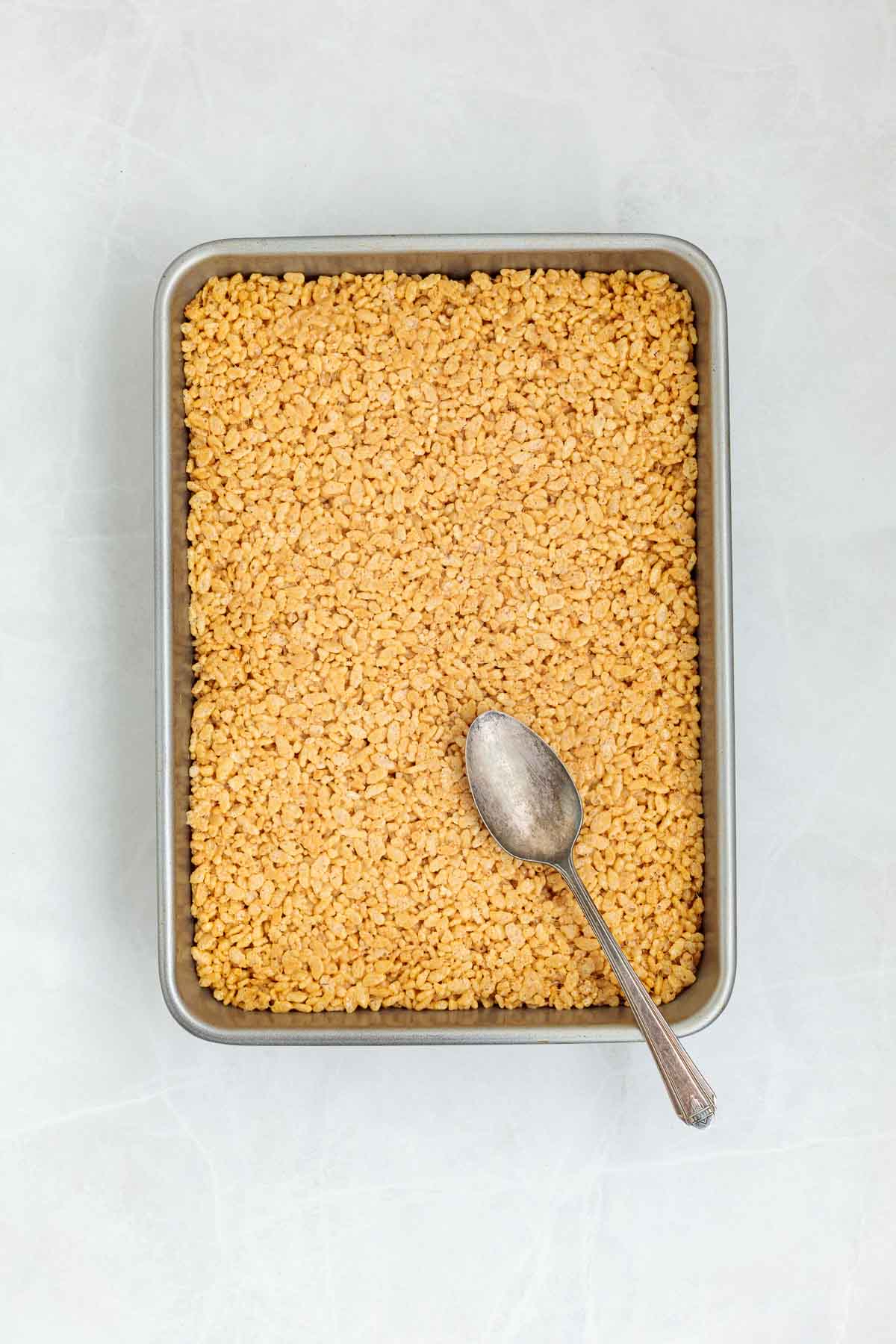 Rice Krispie mixture pressed evenly into a metal baking pan with the back of a spoon resting on top, ready to be topped or cooled.