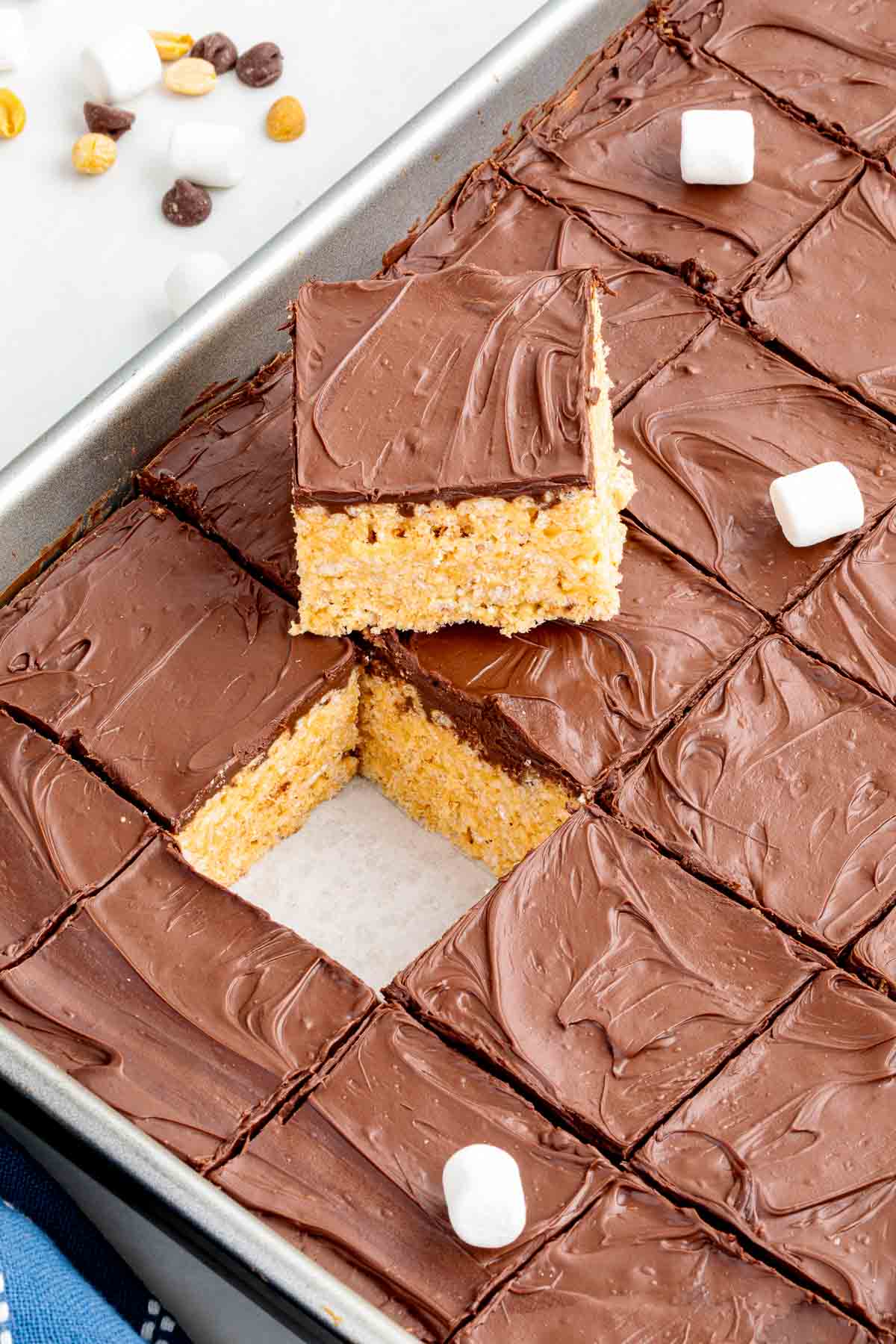 Tray of sliced chocolate-topped Rice Krispie treats with one square lifted out, revealing the crispy peanut butter base underneath.