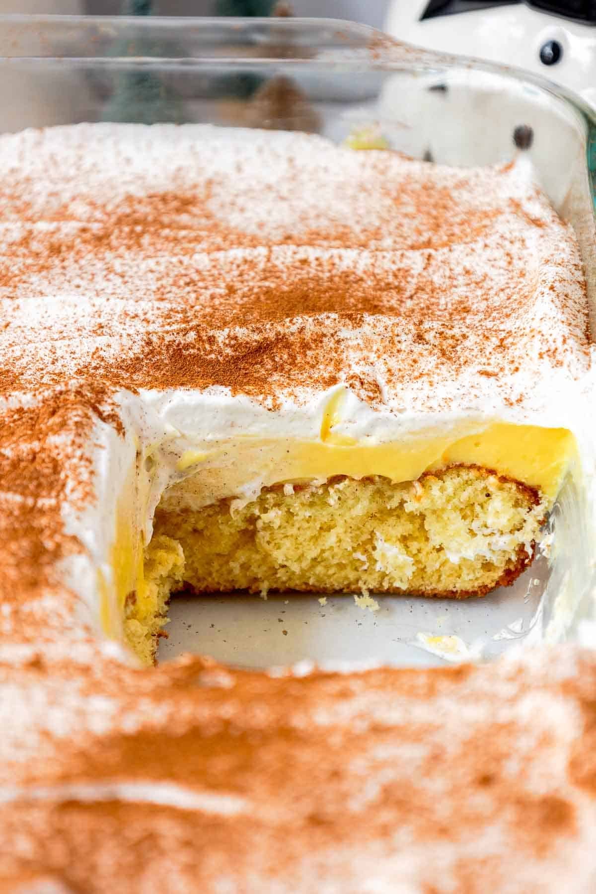 A partially sliced eggnog poke cake in a glass pan, revealing a butter cake layer and creamy eggnog pudding beneath a fluffy cinnamon-dusted whipped topping.