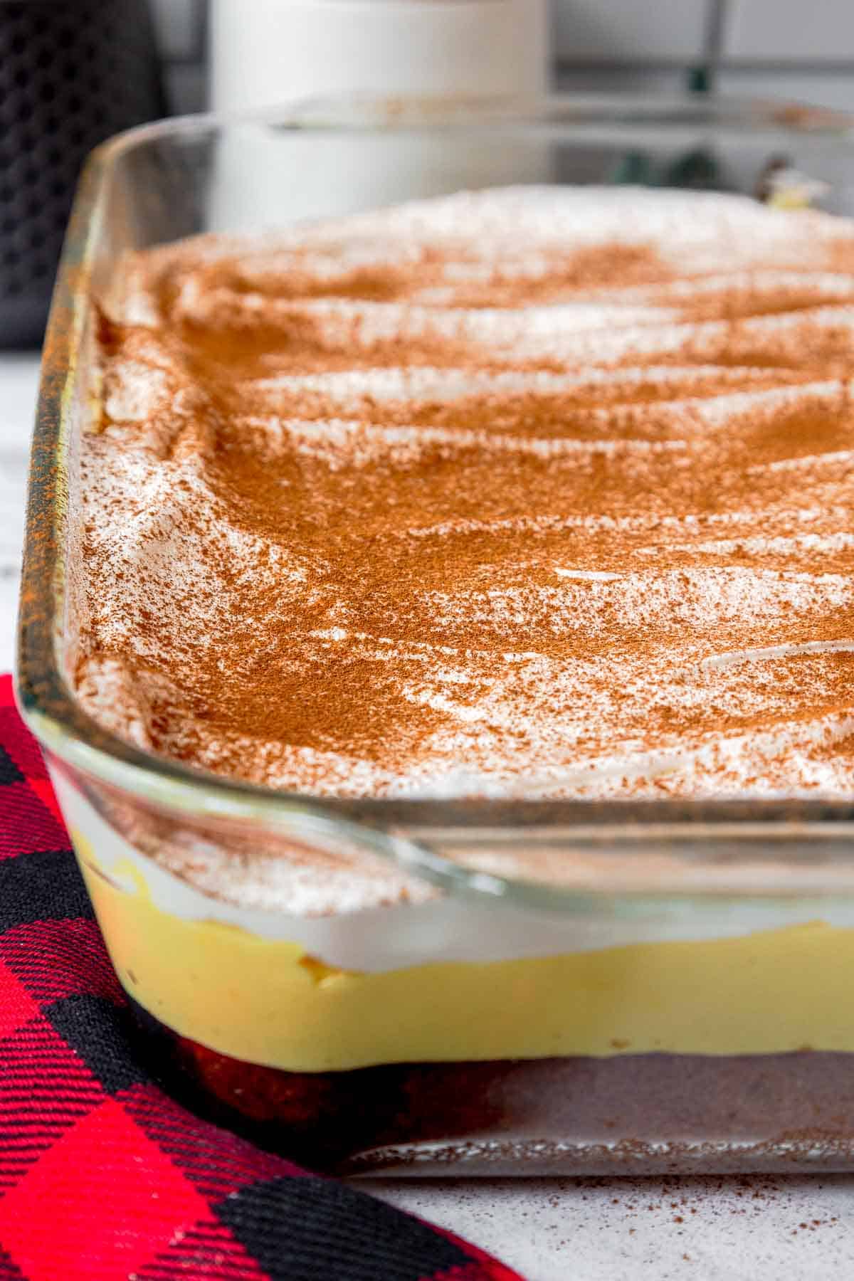 A full, uncut eggnog poke cake in a glass dish with visible layers of butter cake, eggnog filling, and whipped cream sprinkled with cinnamon.