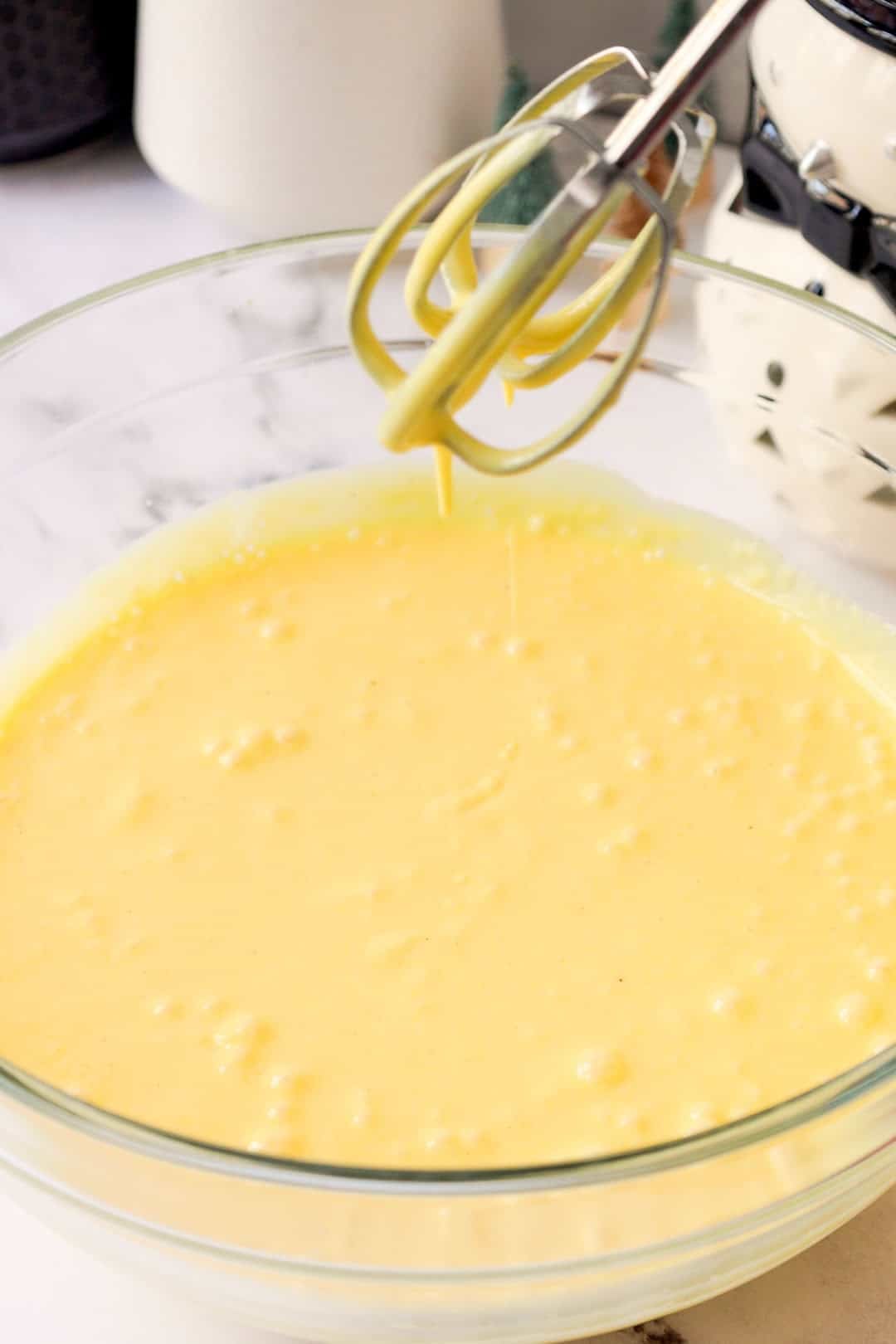 Smooth, yellow eggnog pudding filling being mixed in a bowl with beaters suspended above, showing a loose runny texture.
