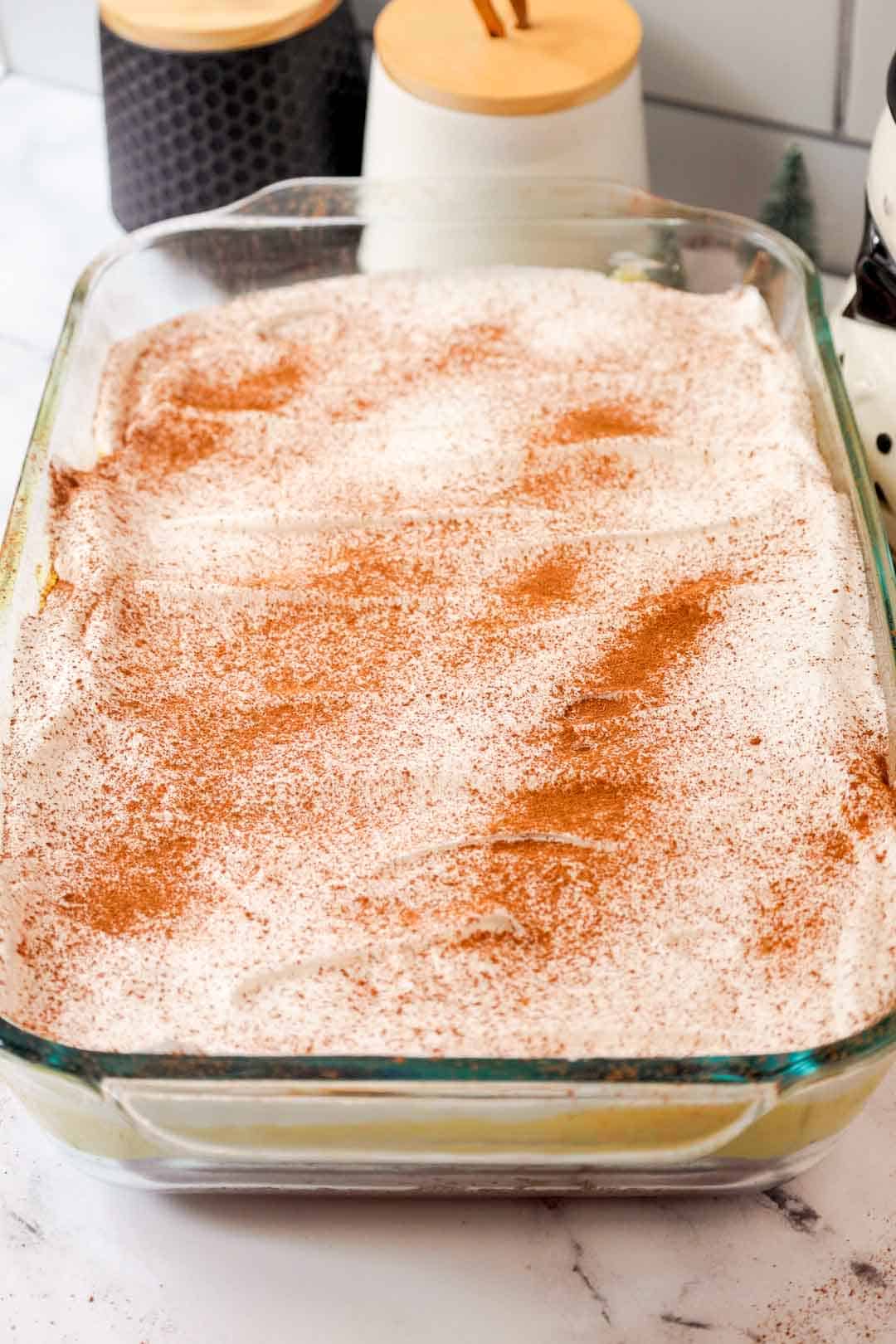 Finished eggnog poke cake topped with whipped cream and a generous dusting of cinnamon and nutmeg, chilled and ready to serve in a glass dish.