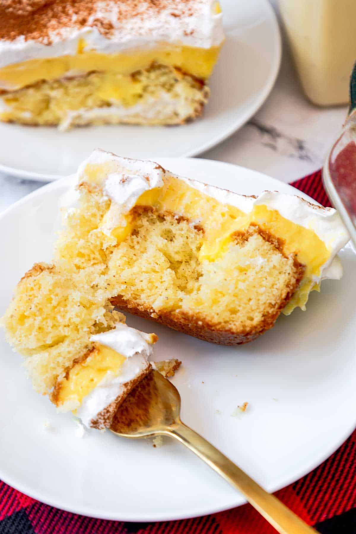 A slice of eggnog poke cake served on a white plate, featuring layers of moist butter cake, creamy eggnog pudding, and whipped topping with a dusting of cinnamon. A fork holds a bite-sized piece showing the fluffy texture and layered filling.
