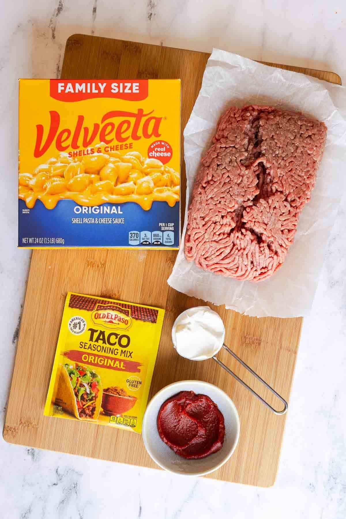 Ingredients for taco mac and cheese laid out on a wooden board, including a box of Velveeta shells and cheese, raw ground beef, taco seasoning packet, tomato paste, and sour cream.