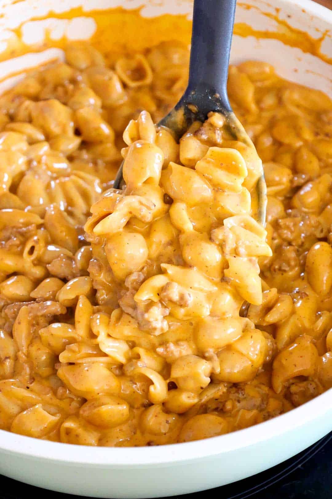 A large spoon lifting a serving of creamy taco mac and cheese out of the pan, showing melted cheese coating the shell pasta and beef.