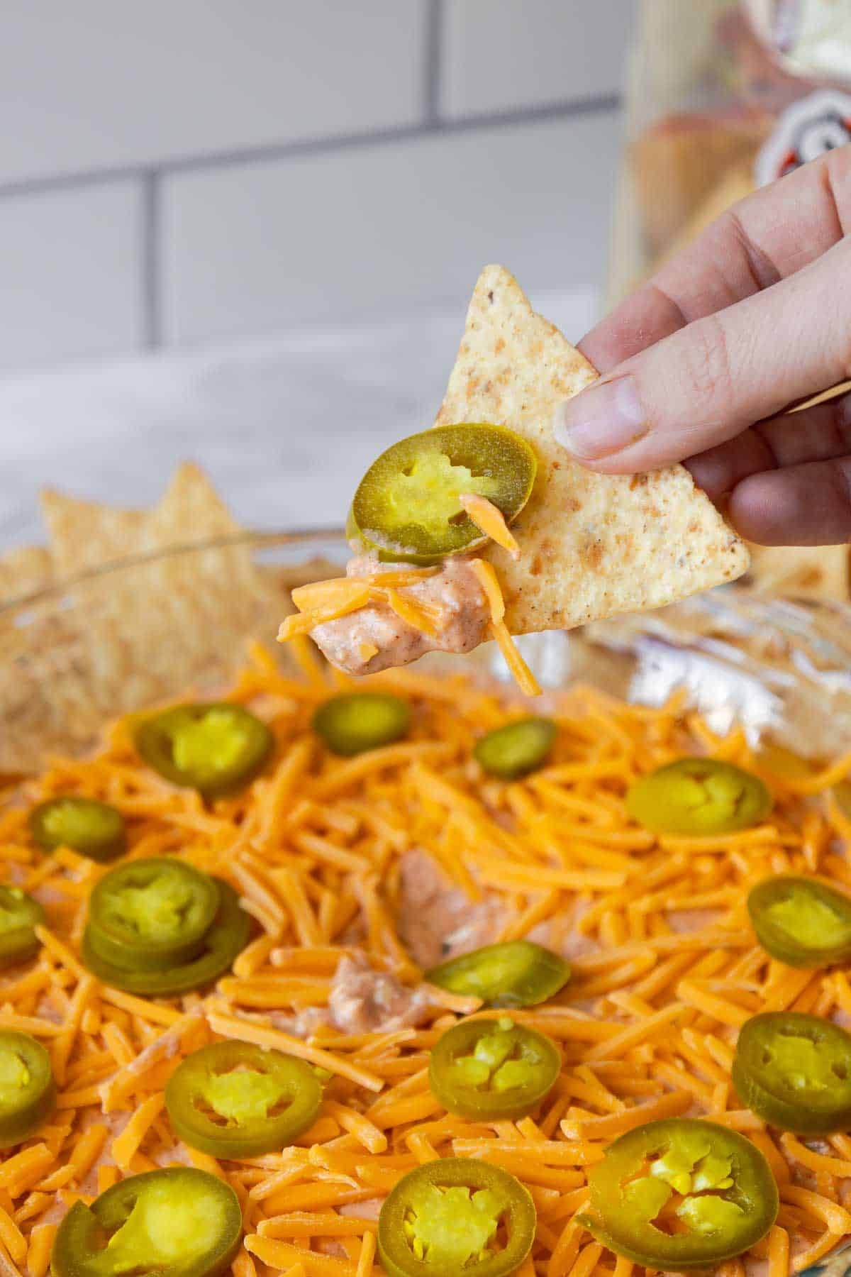A hand holding a tortilla chip scooping up a bite of cream cheese and salsa dip with melted cheese and a jalapeño slice on top.