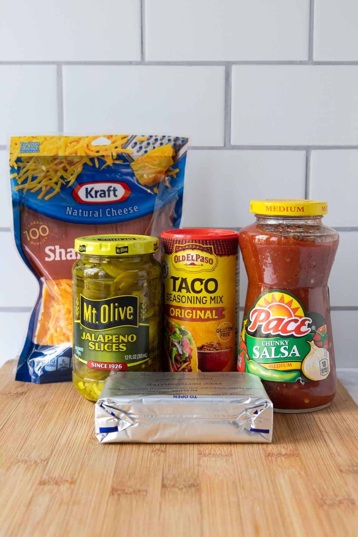 Ingredients for cream cheese and salsa dip arranged on a wooden cutting board, including shredded cheddar cheese, jalapeño slices, taco seasoning, salsa, and a block of cream cheese.