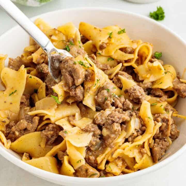 Ground beef egg noodles topped with parsley served in a bowl with a fork lifting cooked noodles and saucy beef.