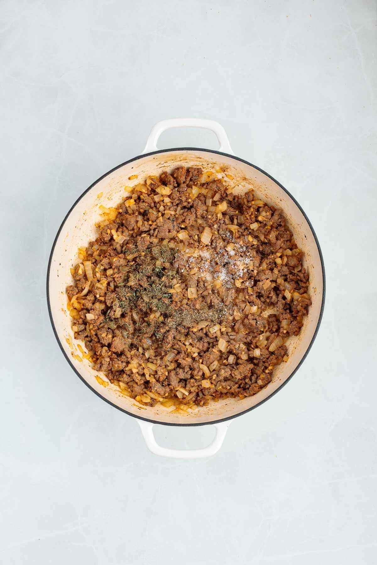 Ground beef and onions cooking in a Dutch oven with dried herbs and seasoning sprinkled over the meat.
