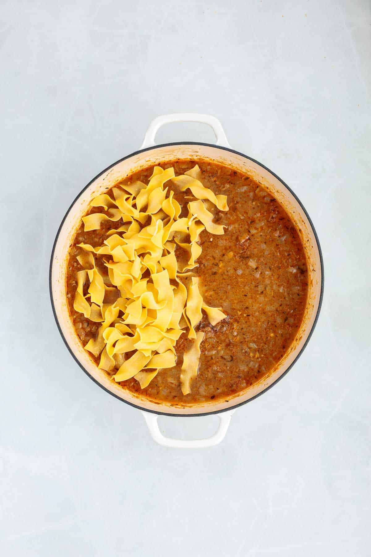 Egg noodles added to a simmering beef sauce in a Dutch oven. The uncooked noodles rest on top of the liquid before being stirred in.