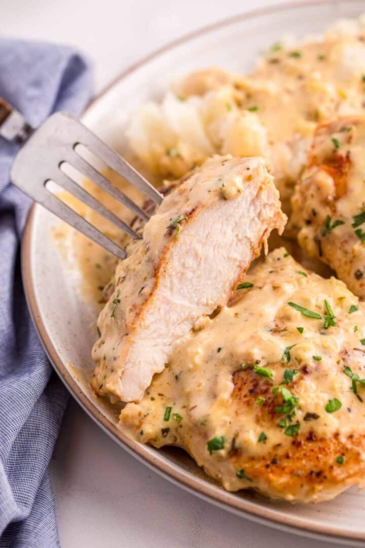 Fork cutting into a chicken breast covered in creamy garlic sauce, showing the inside. Served with mashed potatoes and sprinkled with herbs.