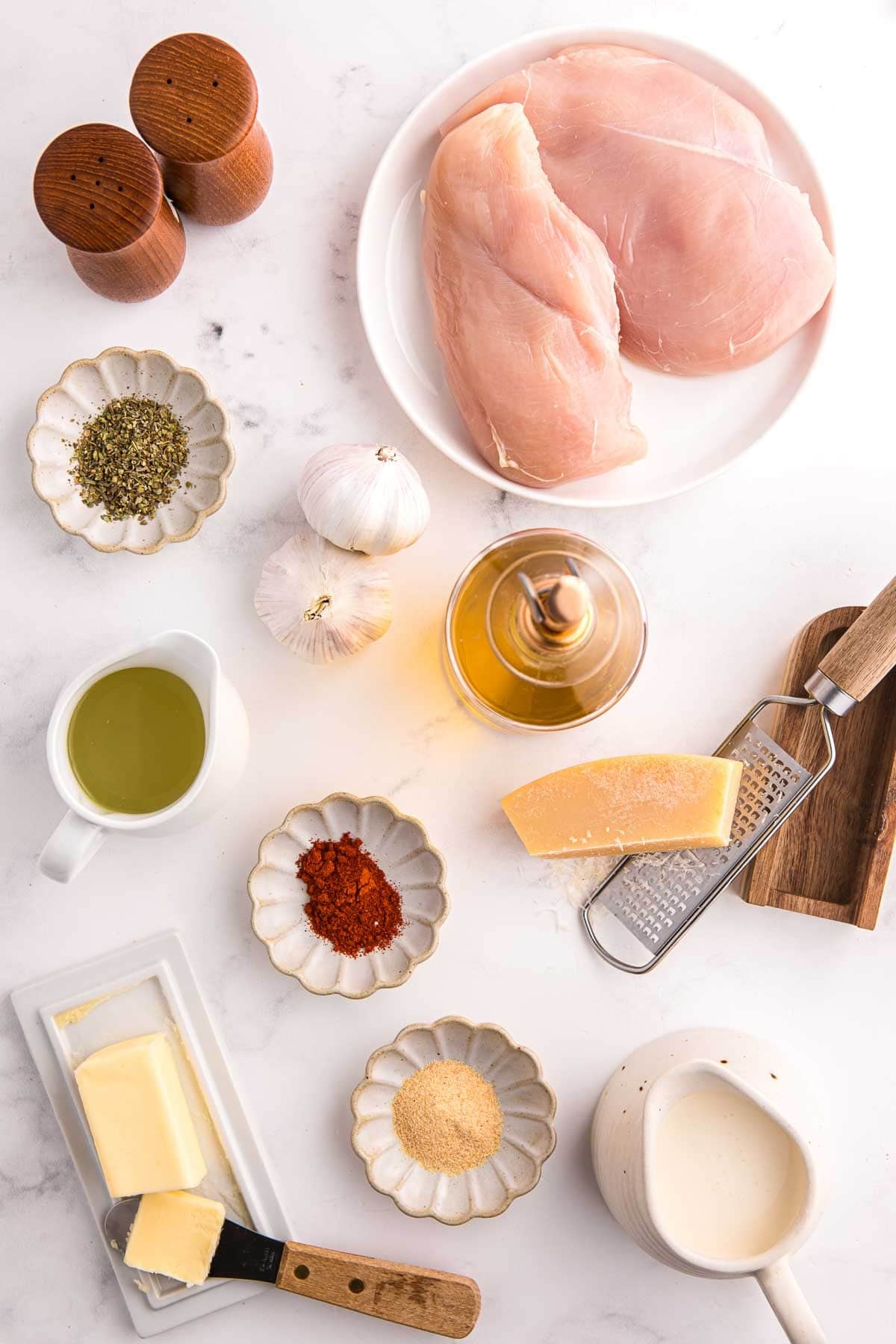 Raw chicken breasts on a plate with garlic, dried herbs, paprika, butter, olive oil, cream, and parmesan cheese. Ingredients laid out to make creamy garlic chicken.