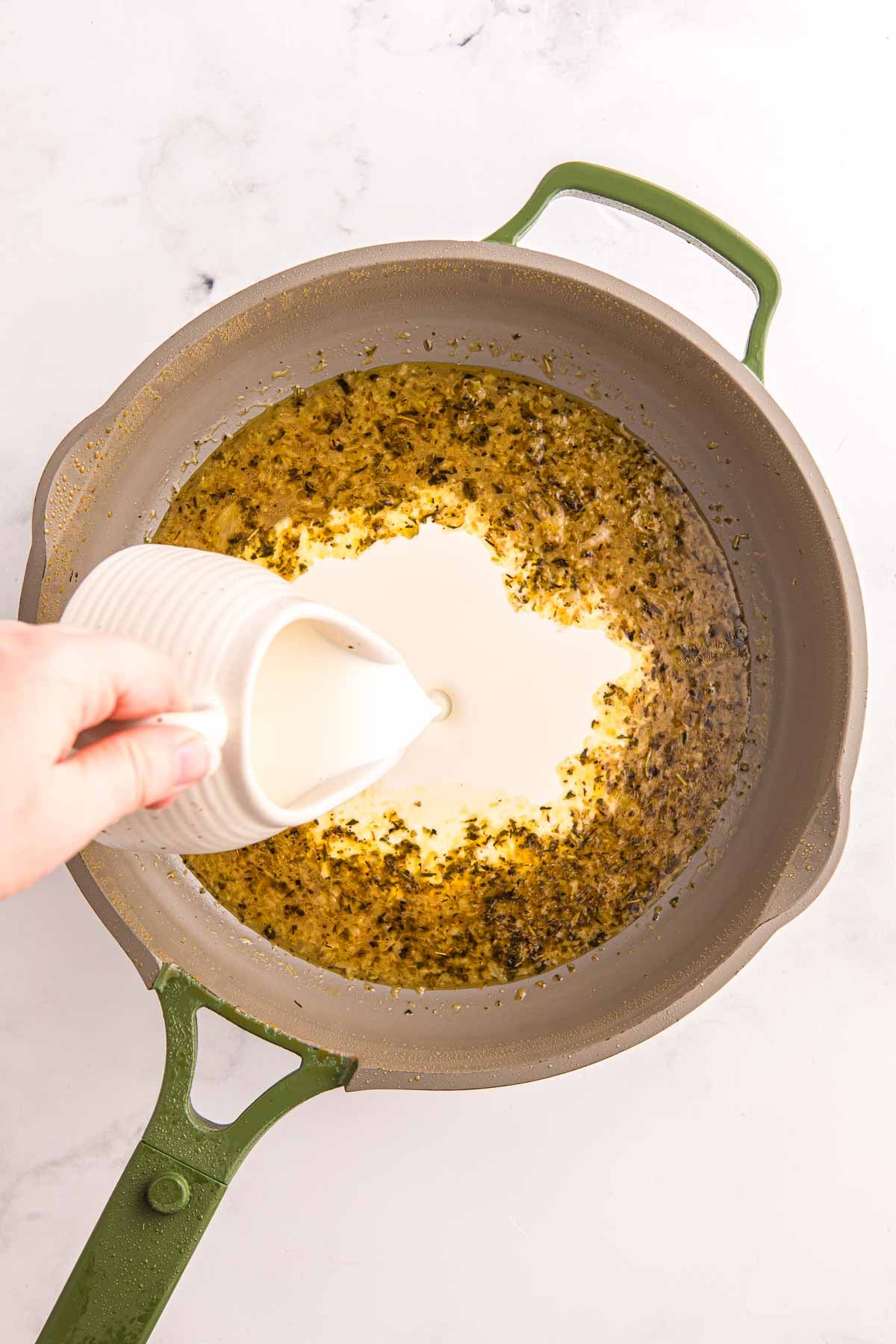 Cream being poured into a skillet with garlic and herbs. Creating the sauce for creamy garlic chicken.