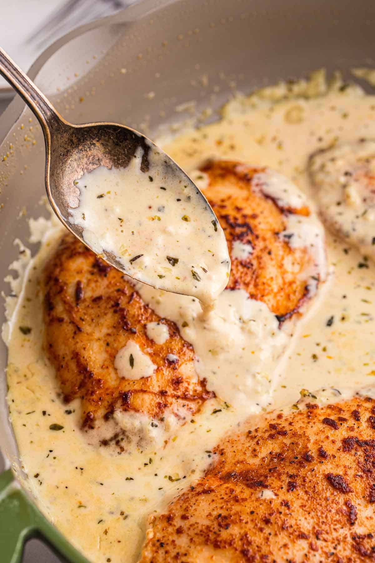 Spoon pouring creamy garlic sauce over browned chicken in a skillet. Finished creamy garlic chicken ready to serve.