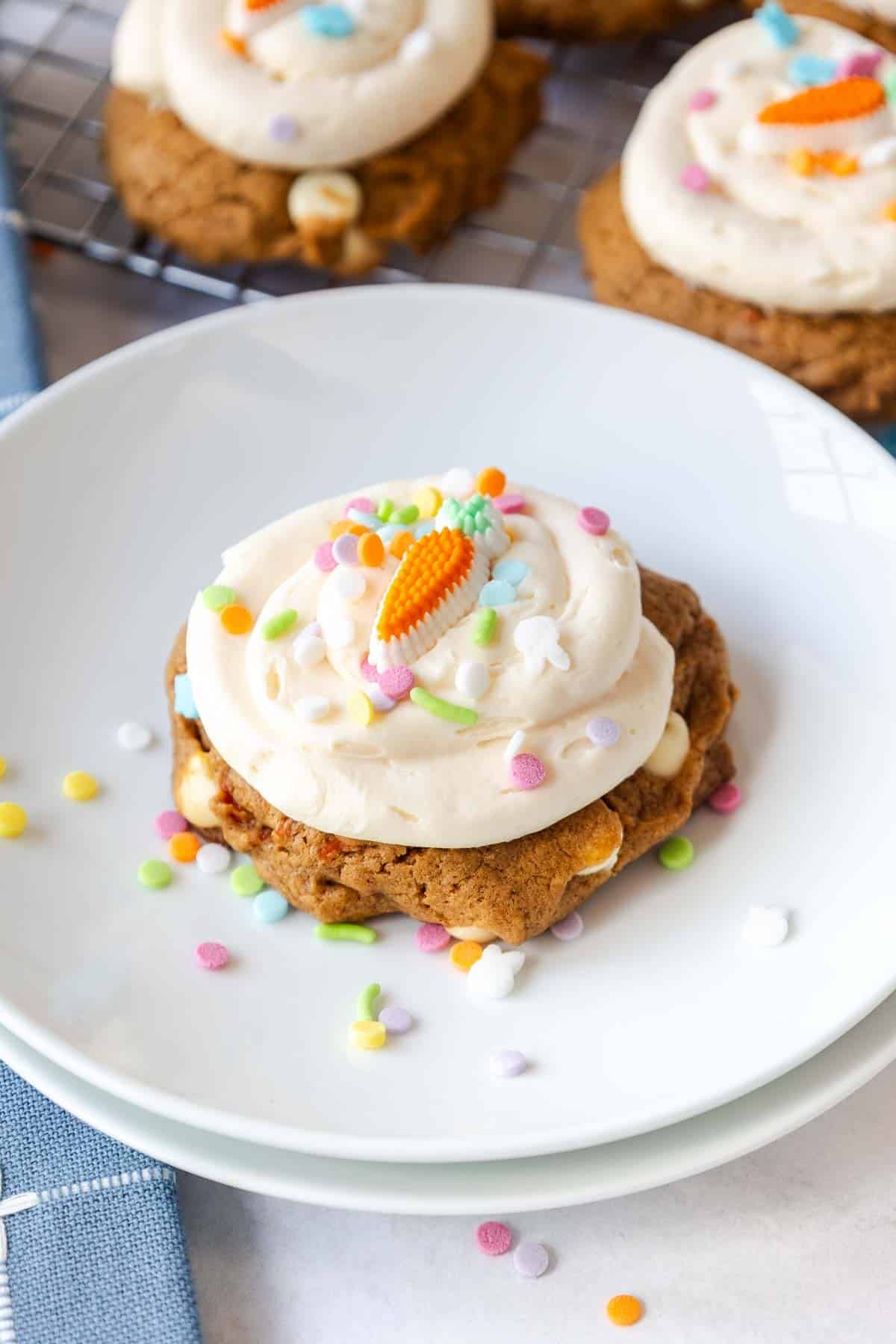 A carrot cake mix cookie topped with cream cheese frosting, colorful sprinkles, and a carrot shaped candy sits on a white plate with extra sprinkles scattered around.