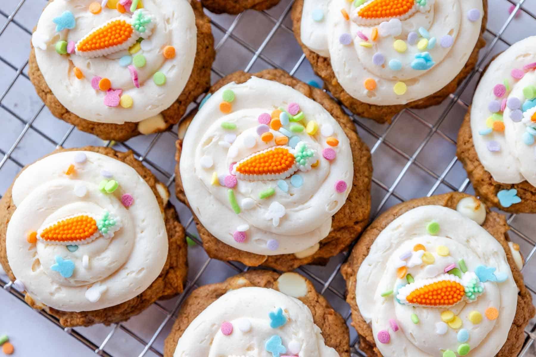 Overhead view of carrot cake mix cookies on a cooling rack topped with swirls of cream cheese frosting, pastel sprinkles, and small carrot shaped candy decorations.