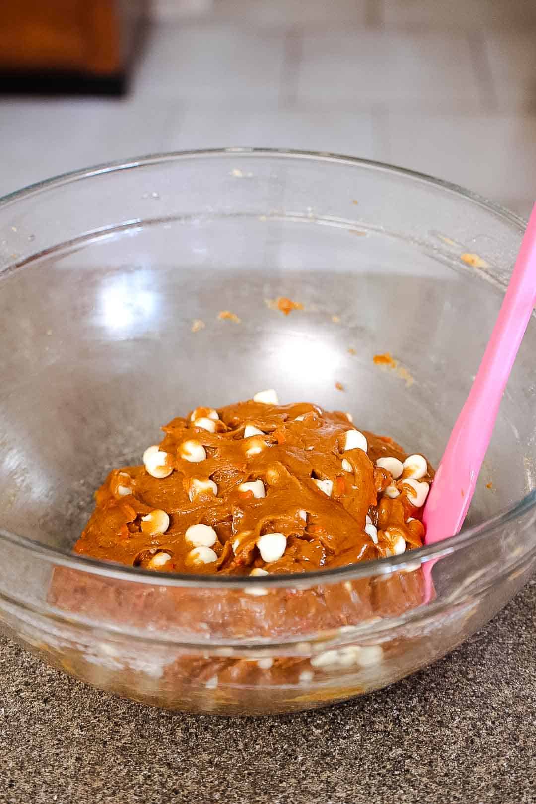 Thick carrot cookie dough with white chocolate chips being mixed in a clear glass bowl with a silicone spatula.