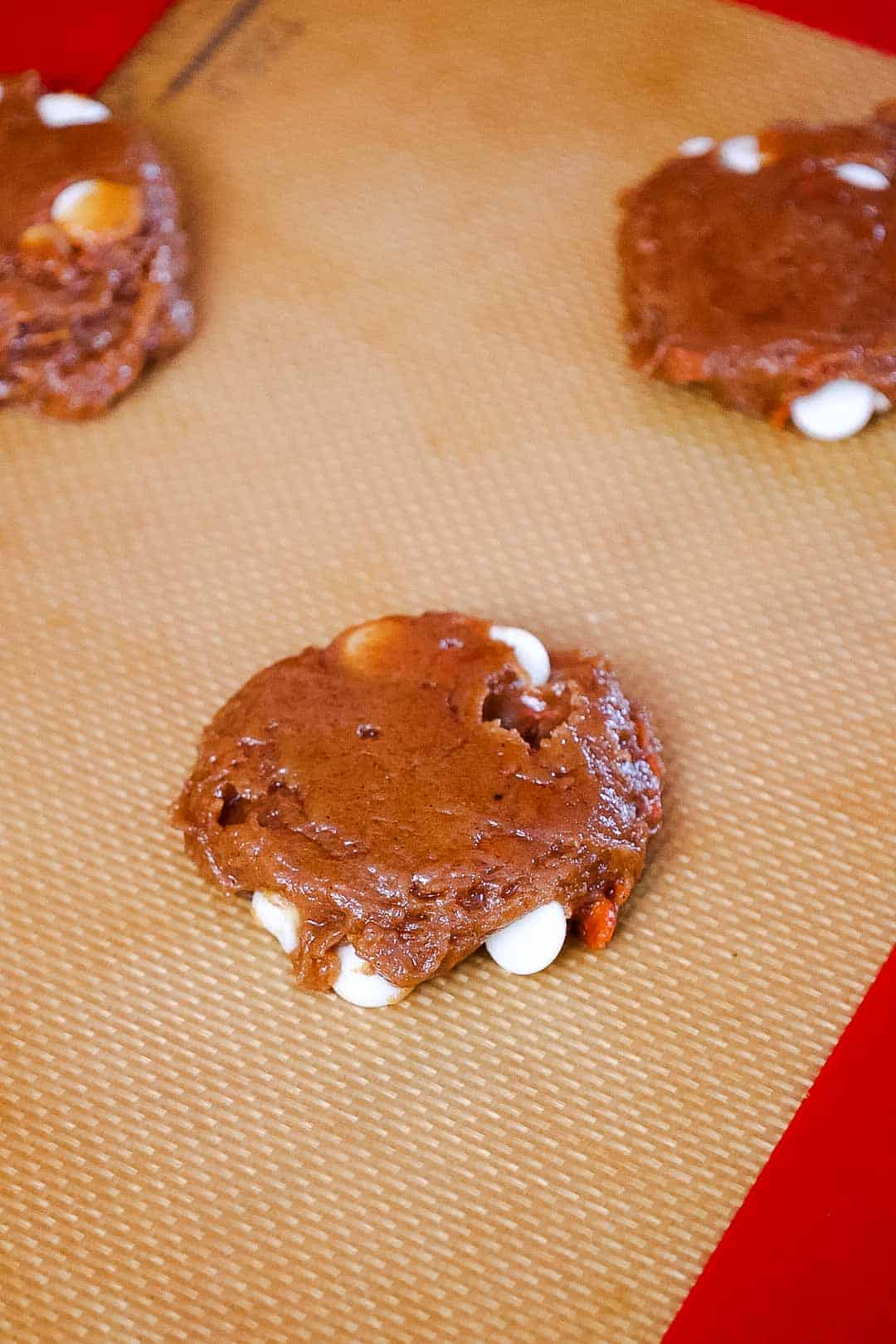Scoop of carrot cookie dough with white chocolate chips flattened onto a silicone baking mat before baking.