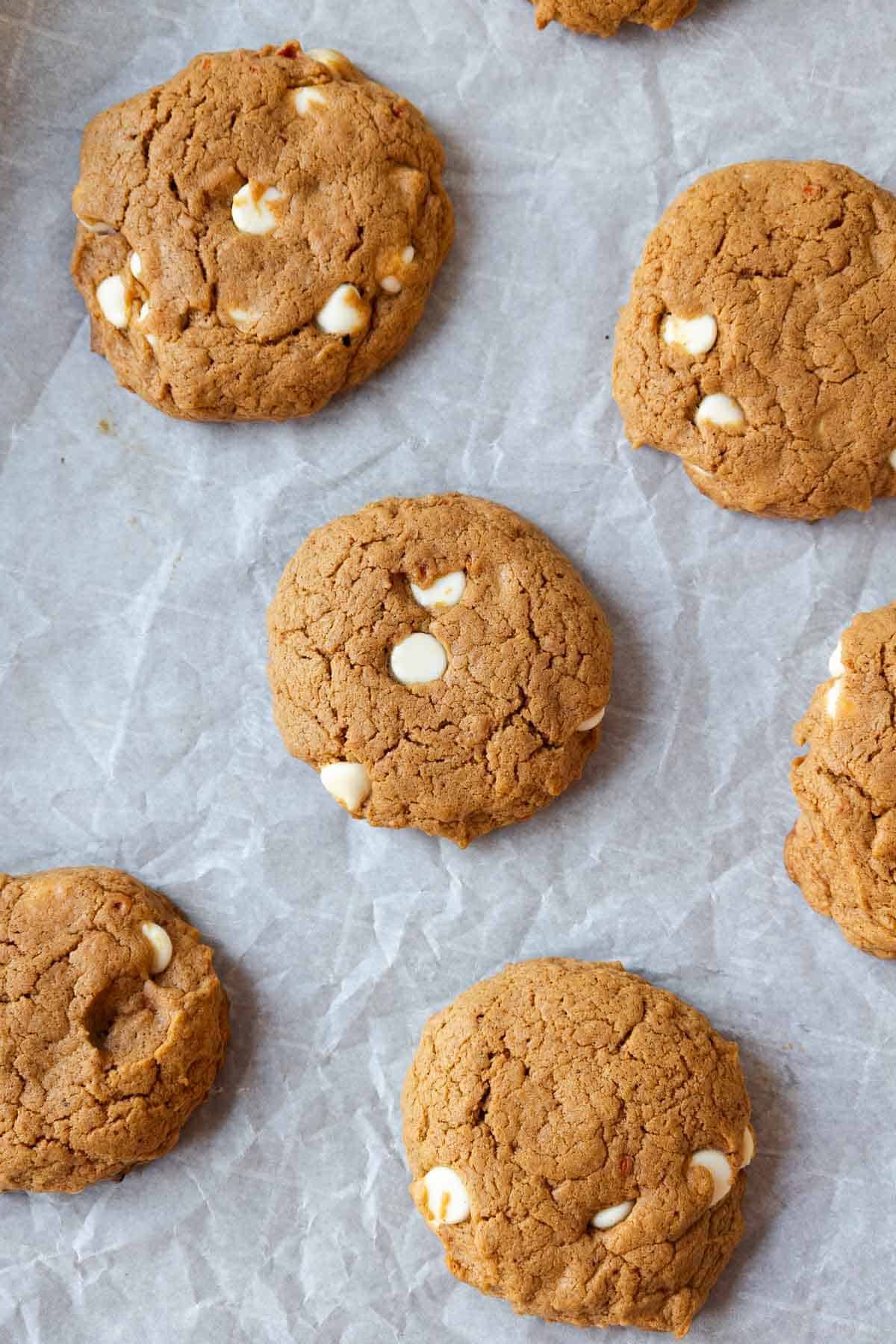 Freshly baked carrot cookies with white chocolate chips spread across parchment paper after baking.