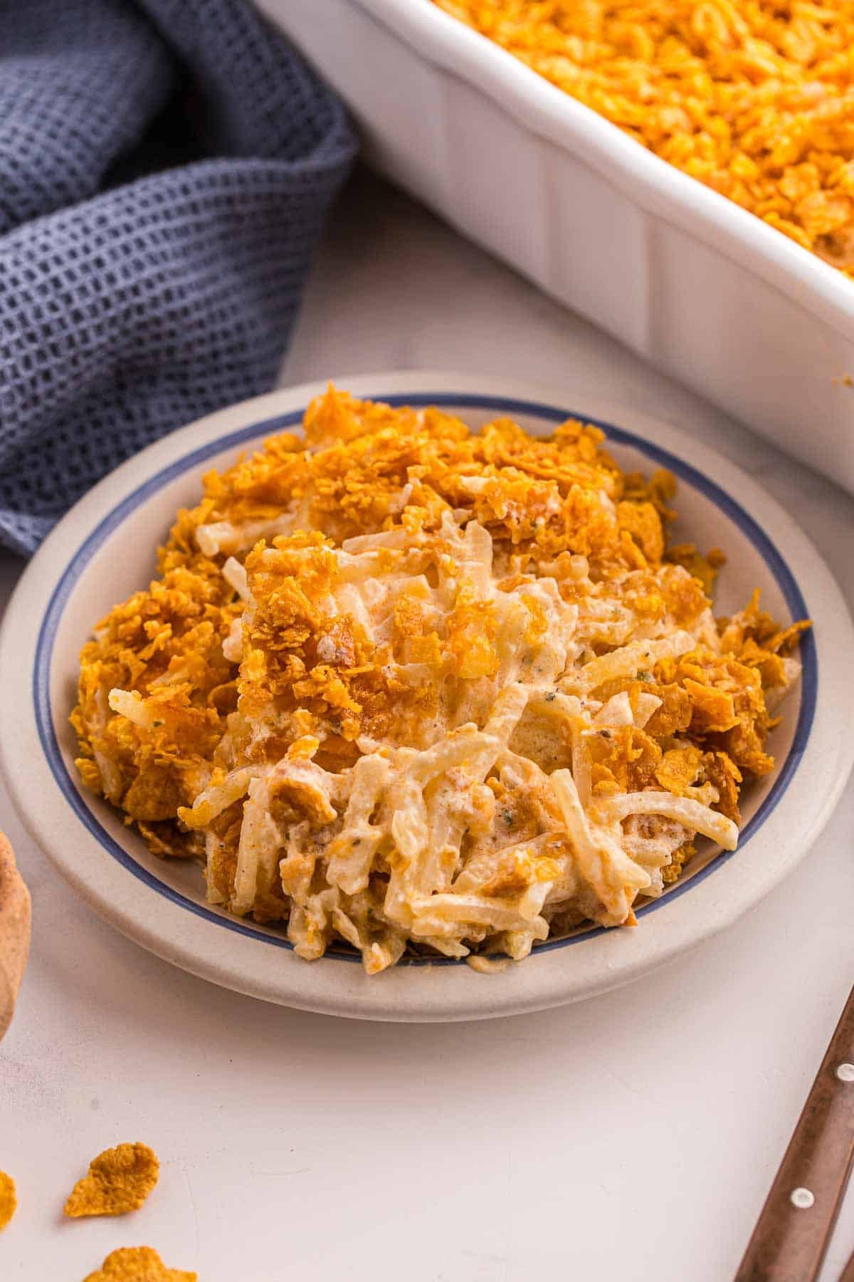 Serving of cheesy hash brown casserole on a plate with a crispy cornflake topping. The dish appears creamy with shredded potatoes and a crunchy top layer.