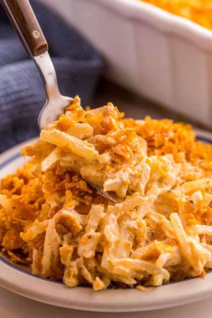 Fork lifting a bite of creamy hash brown casserole mixed with melted cheese and topped with crunchy cornflakes.