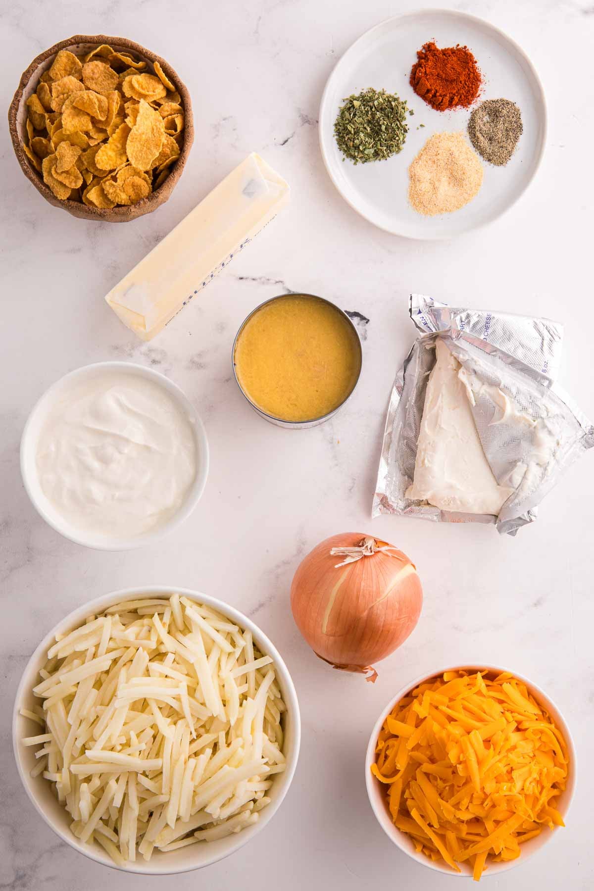 Overhead view of ingredients for hash brown casserole including shredded cheese, sour cream, butter, cream of chicken soup, onion, cornflakes, and spices.