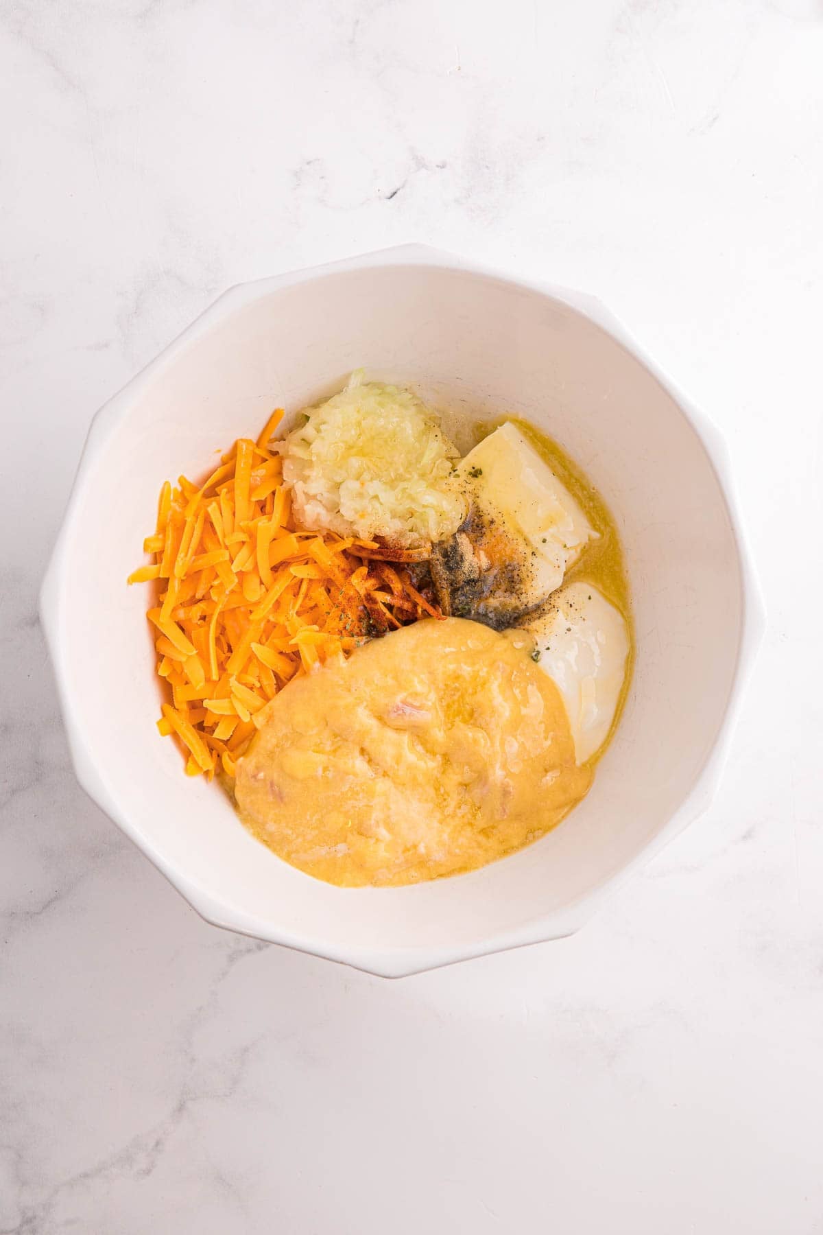 Mixing bowl with ingredients for hash brown casserole including shredded cheese, melted butter, soup, sour cream, chopped onion, and seasonings before being combined.