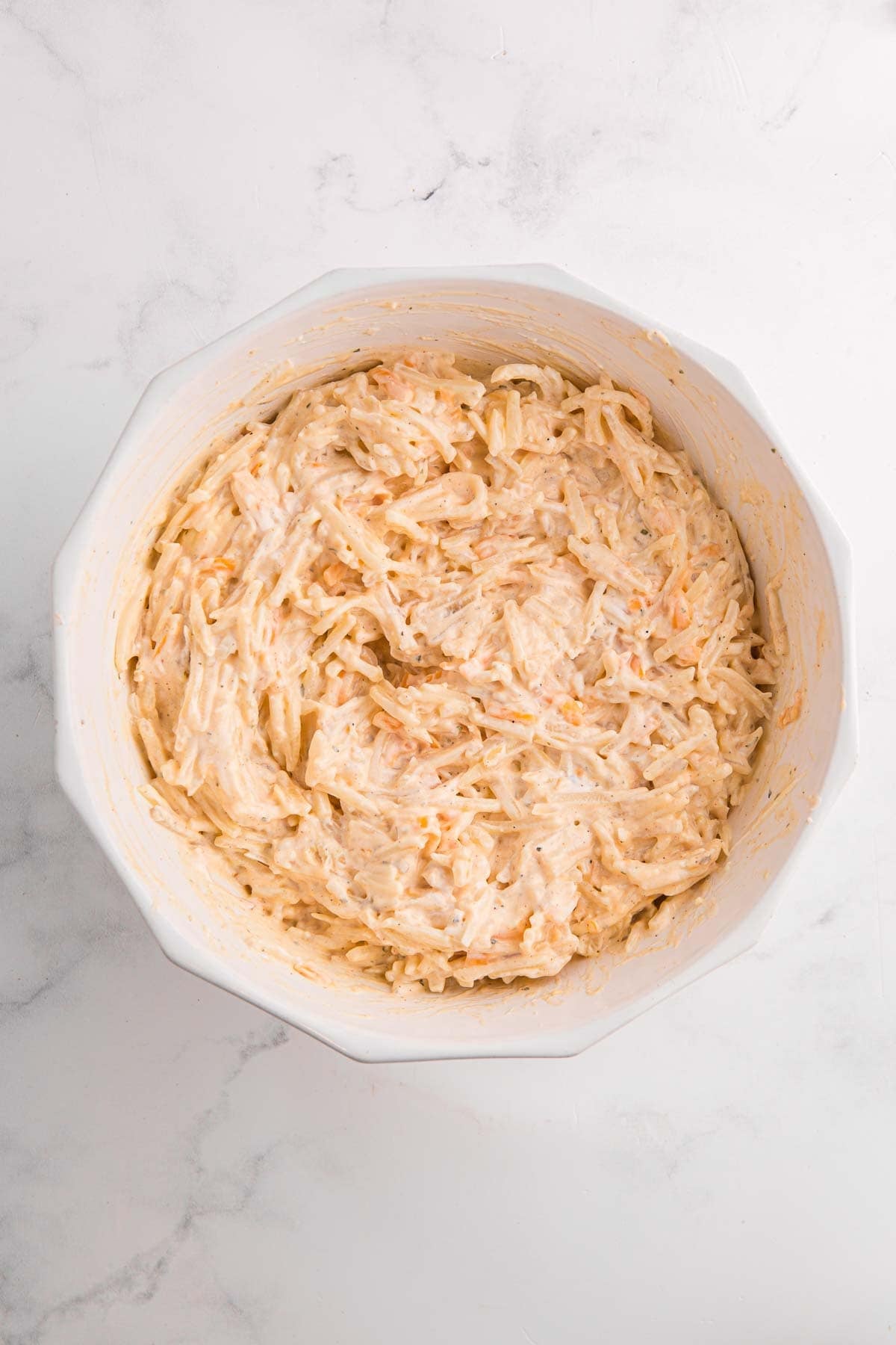 Bowl of fully mixed hash brown casserole filling showing shredded potatoes coated in a creamy cheese sauce with visible seasoning. The mixture looks thick and evenly combined.