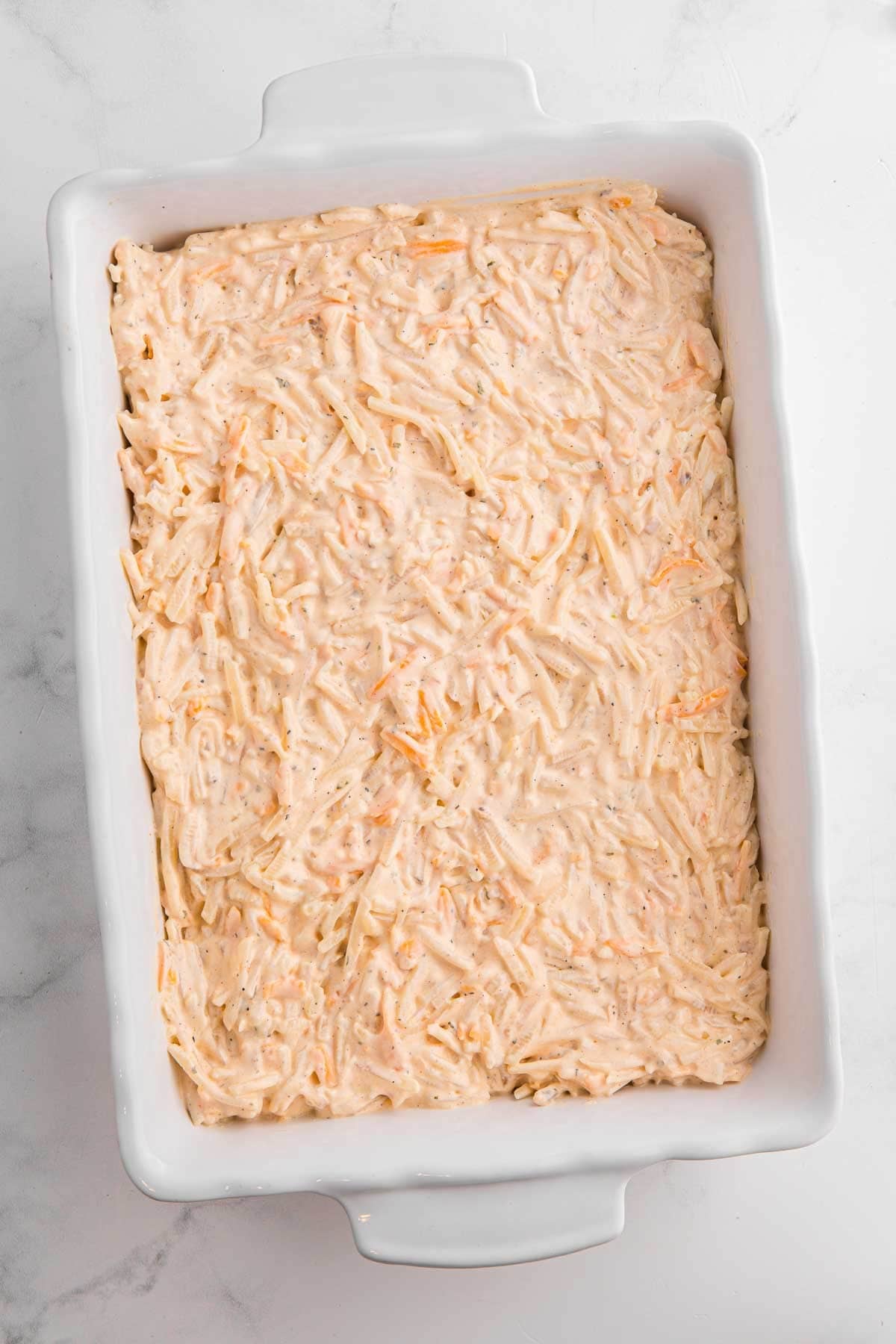 Rectangular baking dish filled with an even layer of uncooked hash brown casserole mixture spread smoothly across the bottom.