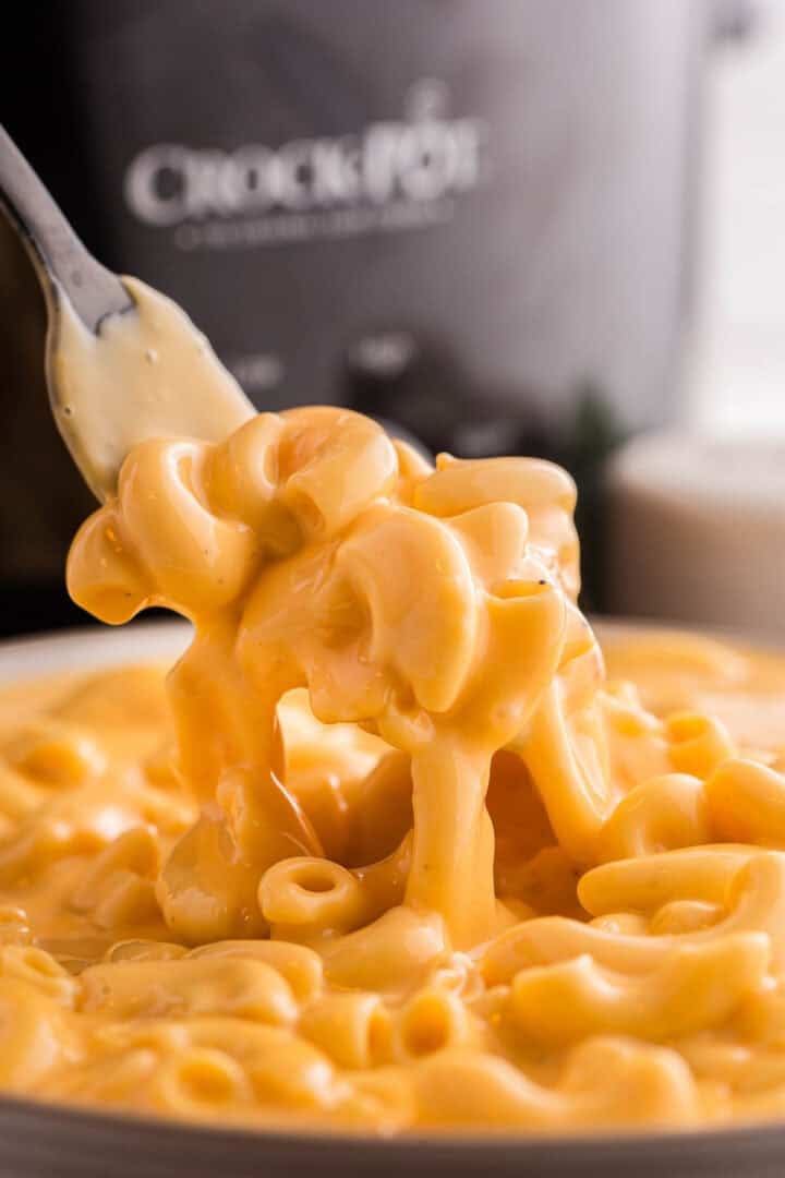 Fork lifting cheese coated elbow macaroni from a bowl of crockpot mac and cheese. The cheese sauce drips back into the dish, highlighting its rich and creamy texture.