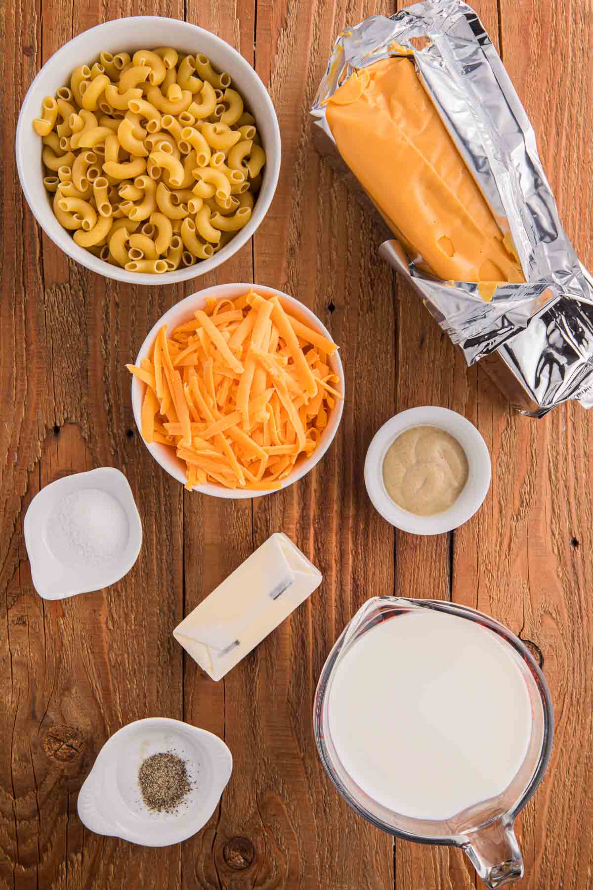 Ingredients for crockpot Velveeta mac and cheese arranged on a wooden surface including elbow macaroni, shredded cheddar cheese, a block of Velveeta, half and half, butter, mustard, salt, garlic powder, and pepper.