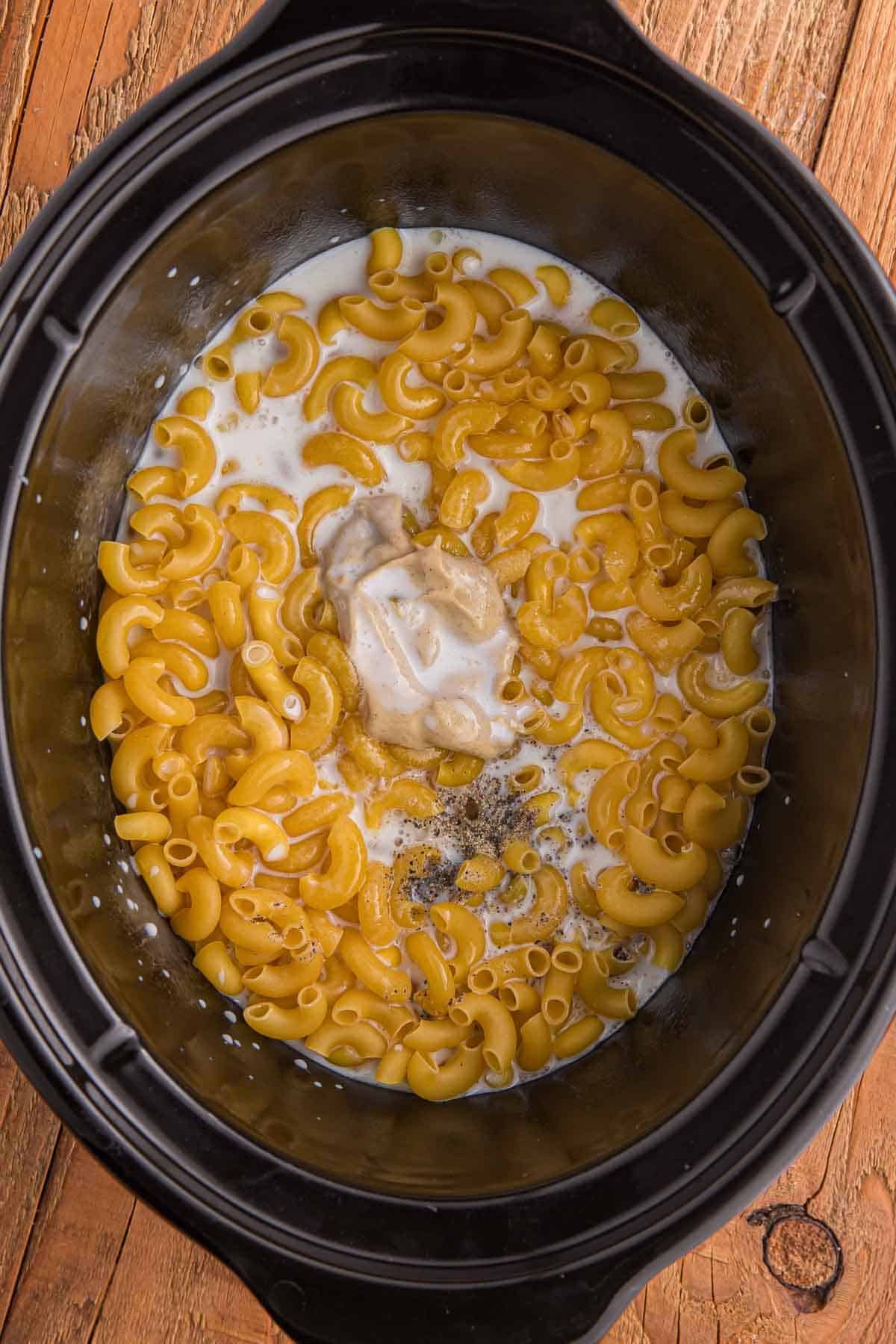 Uncooked elbow macaroni in a slow cooker with half and half, butter, mustard, salt, garlic powder, and pepper added on top.