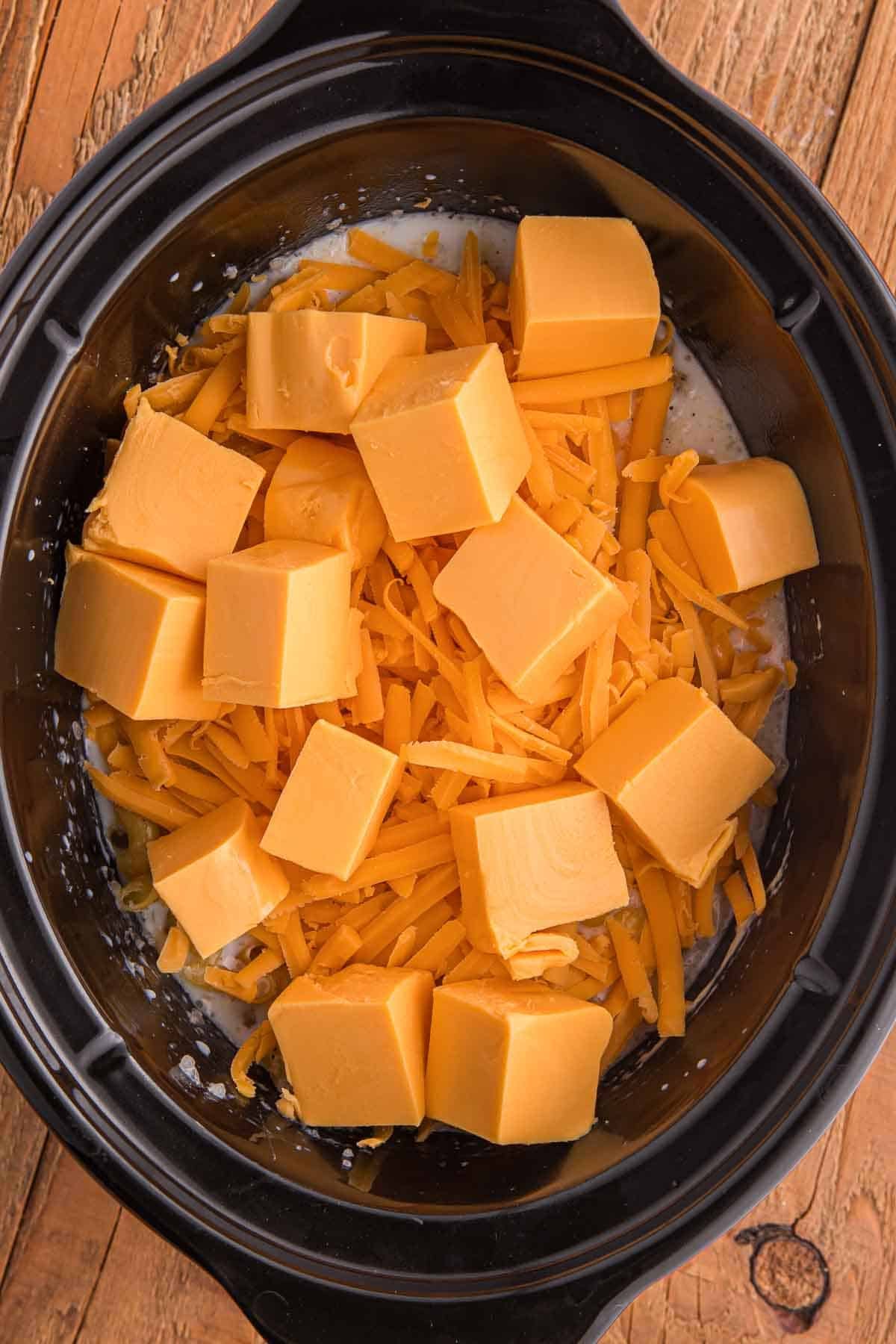 Cubed Velveeta and shredded cheddar cheese added on top of macaroni and milk inside a slow cooker.