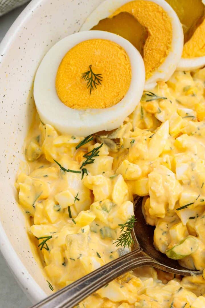 Close up of creamy dill pickle egg salad with chopped eggs, pickles, and fresh dill, topped with sliced hard boiled eggs and served in a white bowl with a spoon.
