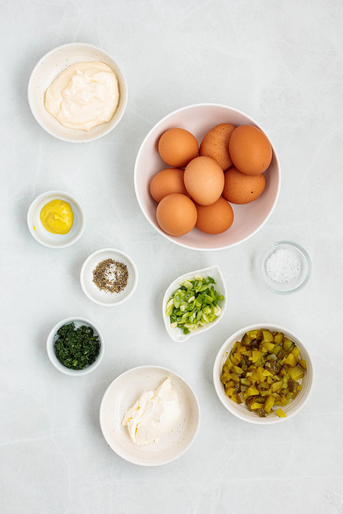 Ingredients for dill pickle egg salad arranged on a countertop including whole eggs, mayonnaise, cream cheese, mustard, chopped pickles, green onions, dill, salt, and pepper.