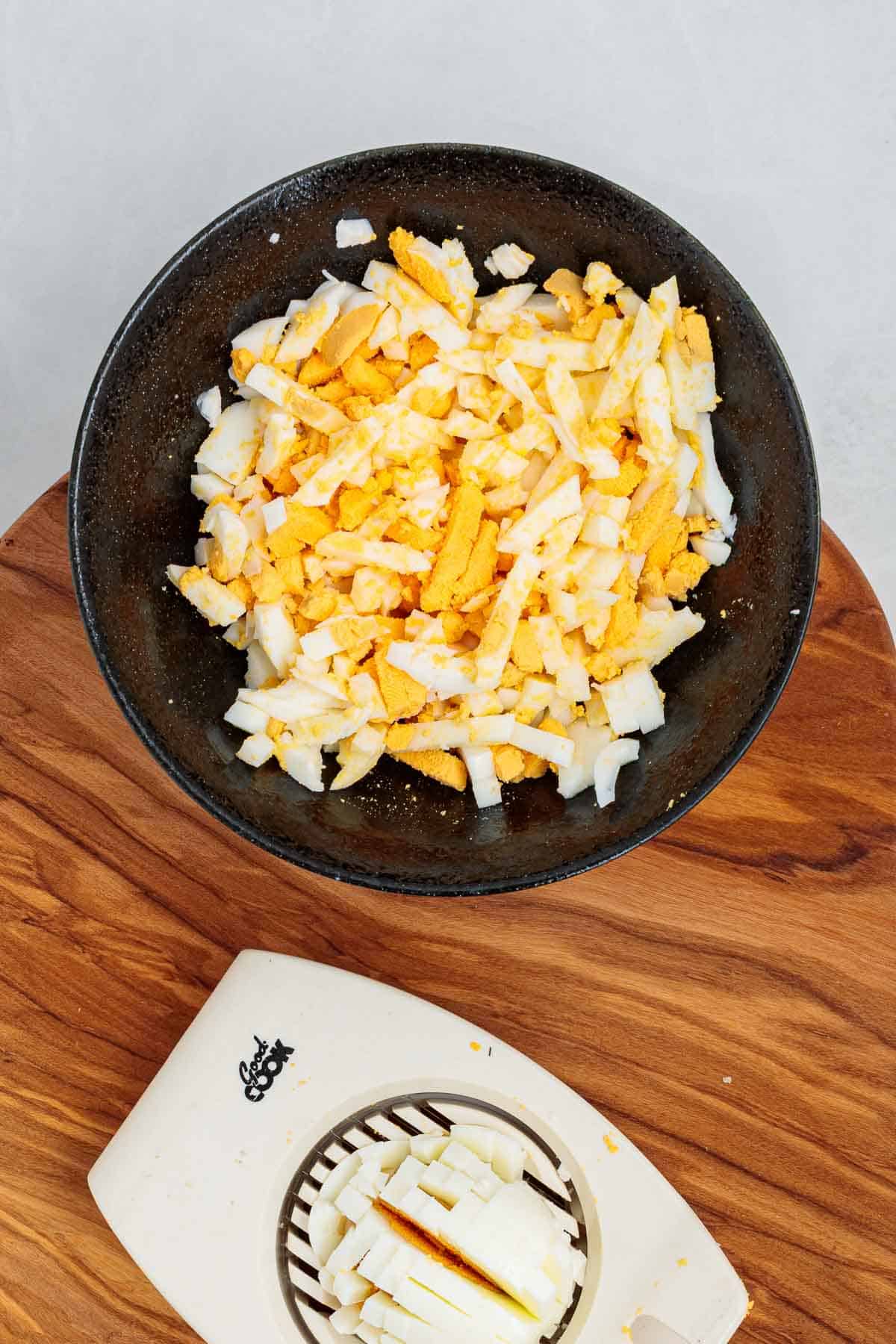 Chopped hard boiled eggs in a bowl with visible yolk and whites, with an egg slicer on a wooden cutting board showing part of the prep process.