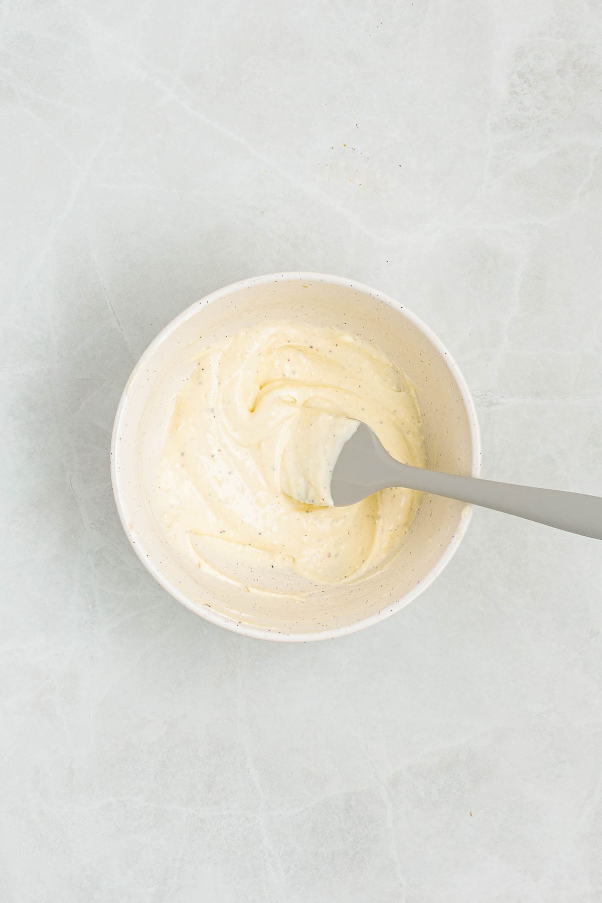 Mixing bowl of smooth creamy dressing made with mayonnaise, whipped cream cheese, dijon, and seasonings, being mixed with a spatula on a light countertop.