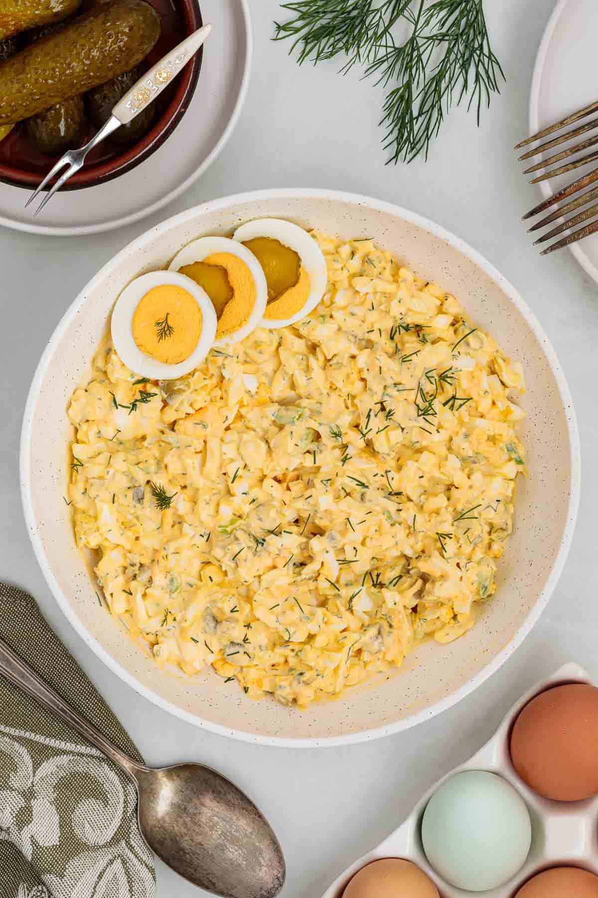 Large serving bowl of dill pickle egg salad garnished with sliced hard boiled eggs and fresh dill, surrounded by pickles, utensils, and whole eggs on a table.