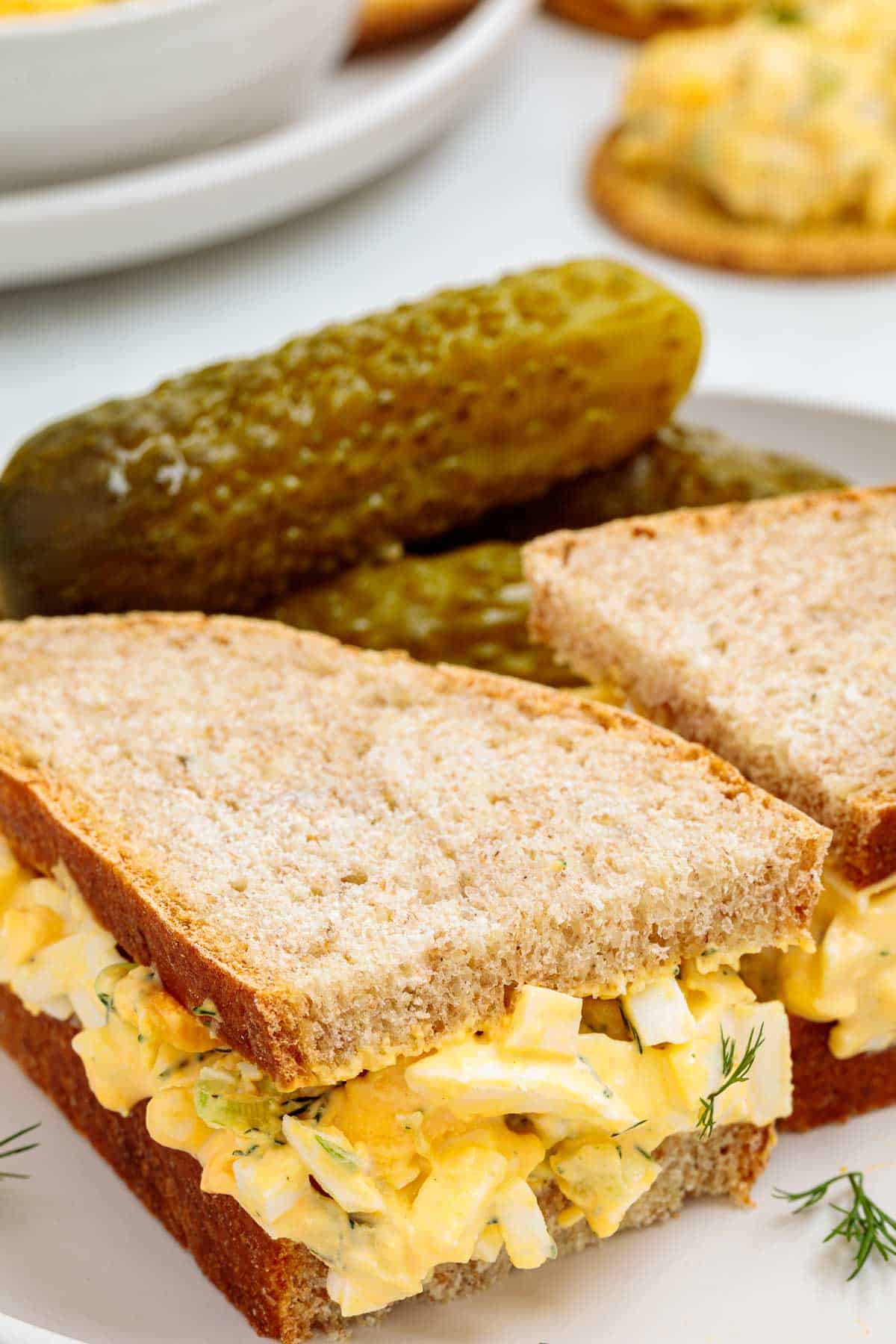 Dill pickle egg salad sandwich made with soft sliced bread filled with creamy egg salad, served with whole pickles on the side.